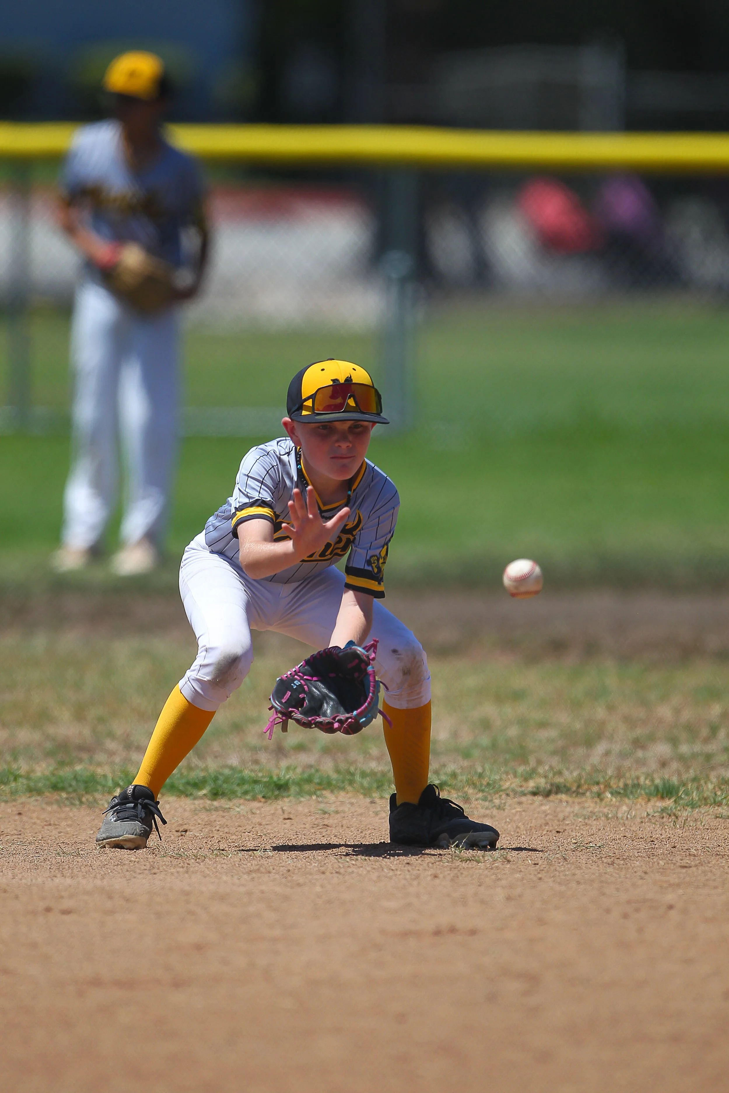 A young baseball player in a gray and yellow uniform is crouching on the dirt infield, preparing to field a hard hit ground ball. The baseball is midair near the glove on his left hand.