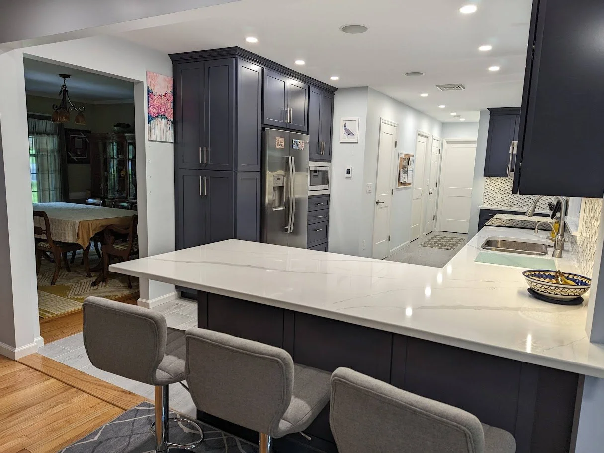 Kitchen with wooden cabinets, white countertop, black stovetop, built-in oven, stainless steel refrigerator, stainless steel sink, and large windows overlooking an outside porch and green yard.