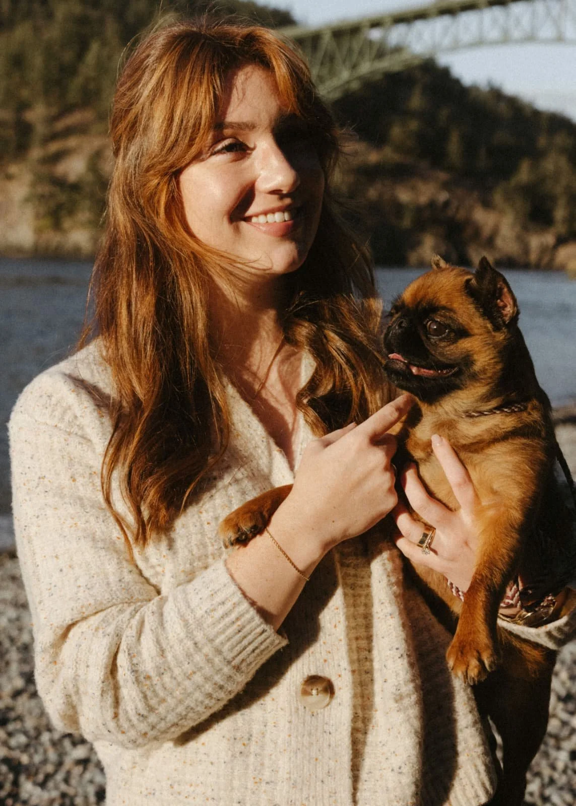 A woman with reddish hair smiling while holding a small brown and black dog near a body of water with a bridge in the background.