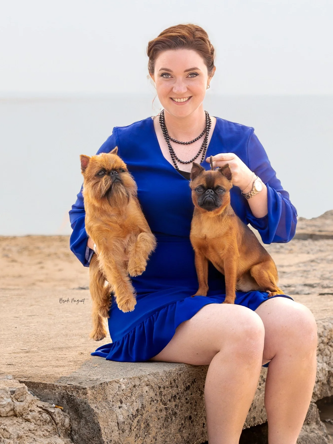 A woman in a blue dress sitting outdoors on a rock holding two small dogs, one in each arm, near a body of water.