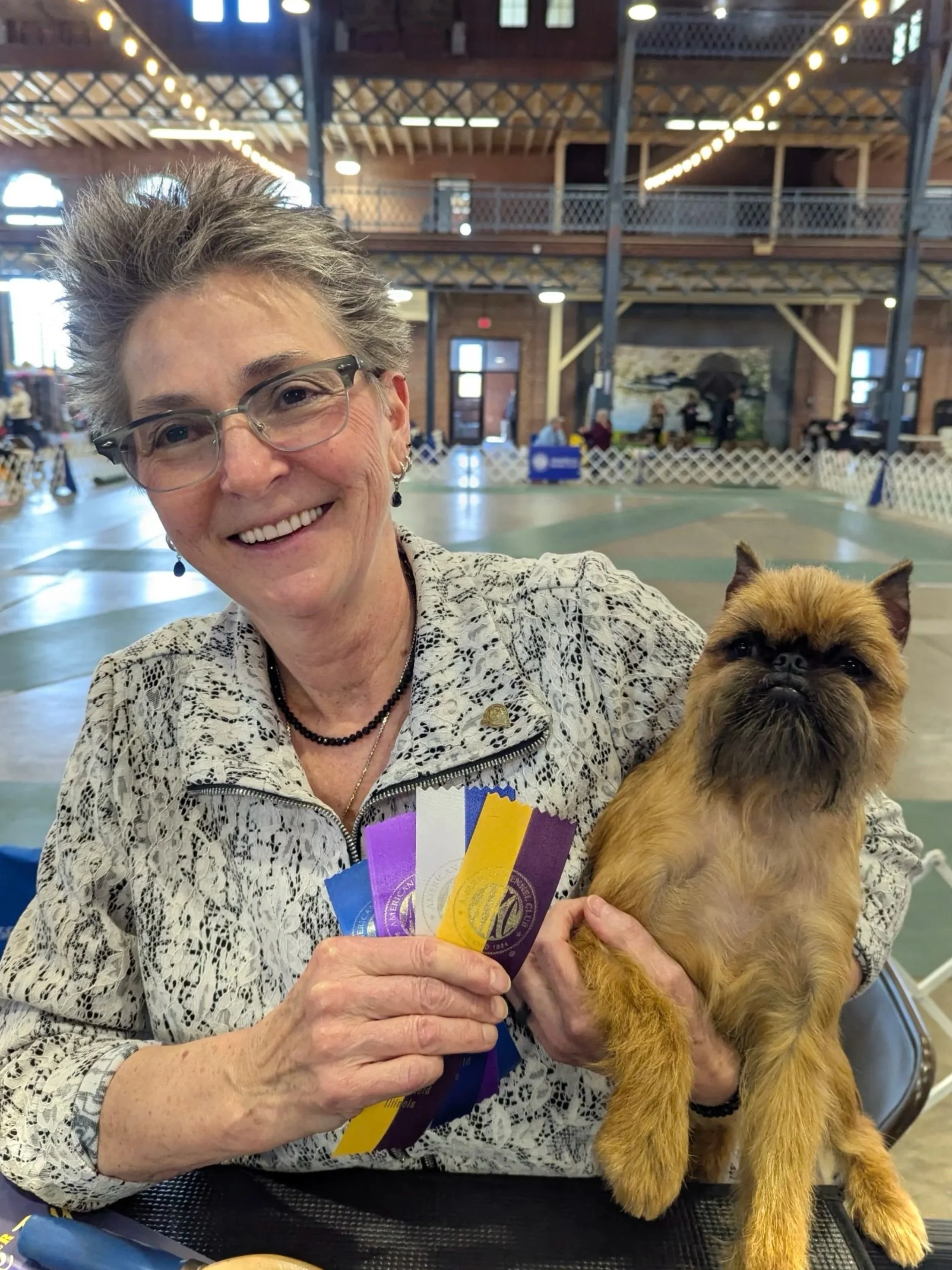 A smiling woman with short gray hair, glasses, and a patterned jacket holding ribbons, sitting indoors in a large wooden building. She is holding a small brown dog with a flat face, possibly a Brussels Griffon, in her lap.