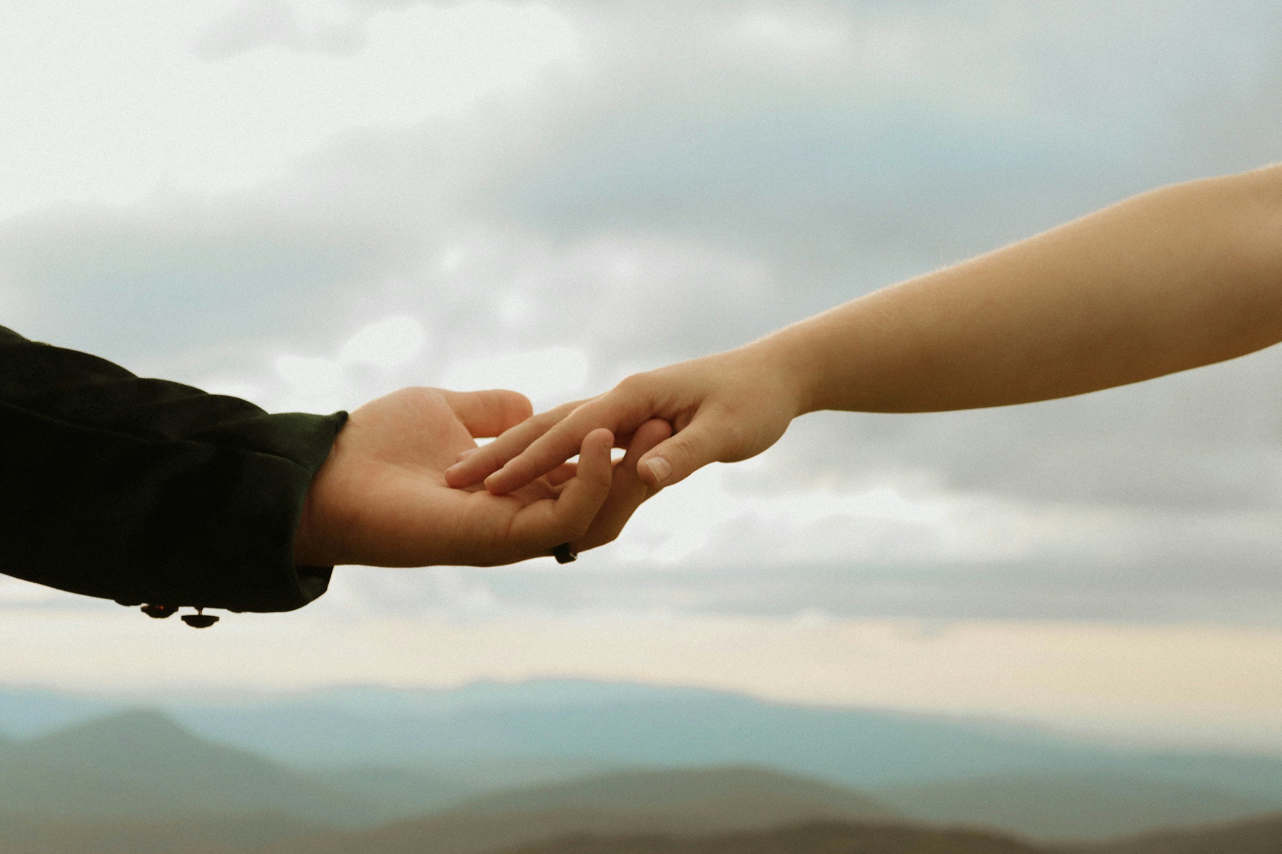 Two hands reaching out and touching each other, one in a dark jacket and the other in a light skin tone, with a cloudy sky in the background.