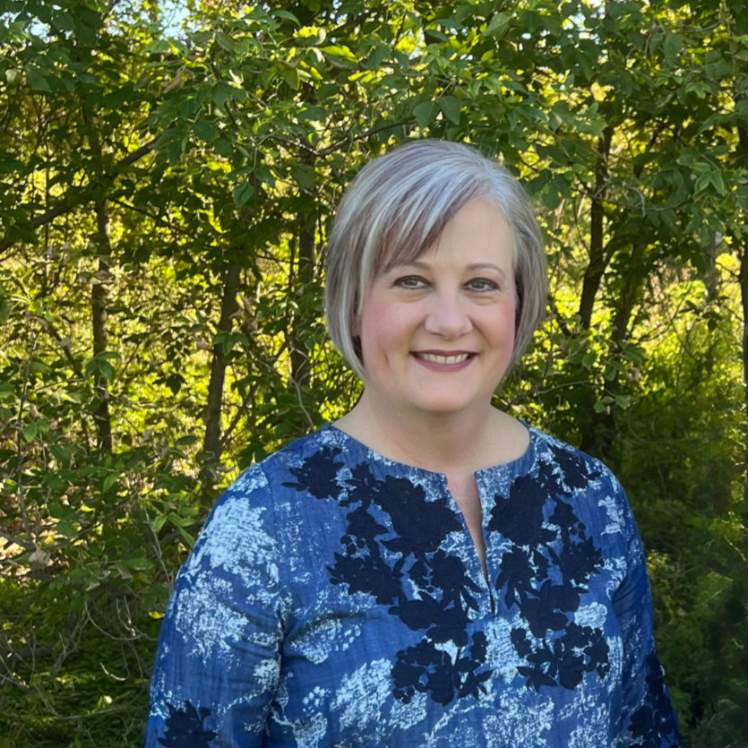 The owner, Lynn Misirian, with short gray hair smiling outdoors, wearing a blue patterned top, standing in front of green foliage.