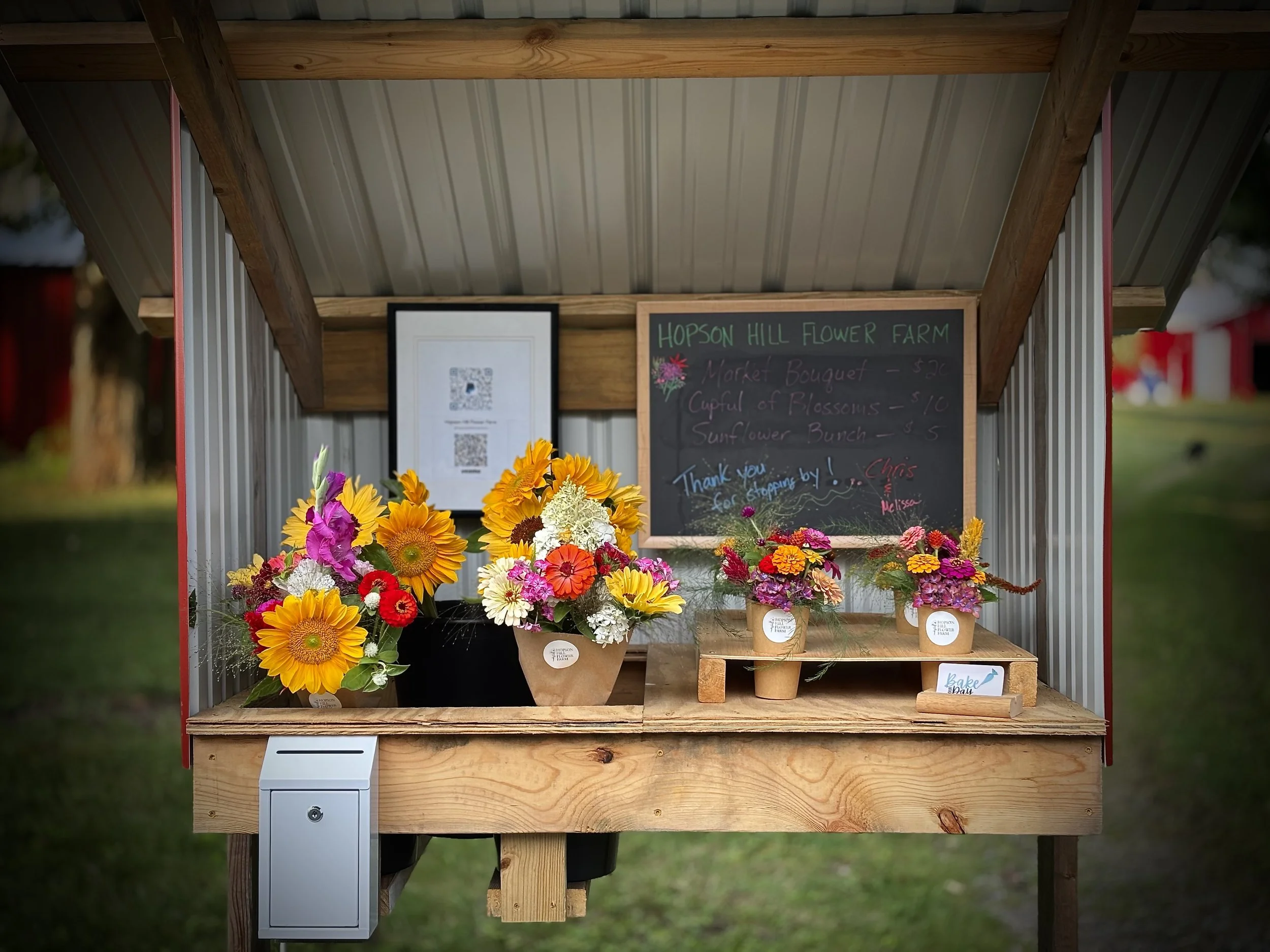 Flower stand with colorful bouquets, handwritten chalkboard sign displaying flower prices, and QR codes on the back wall.