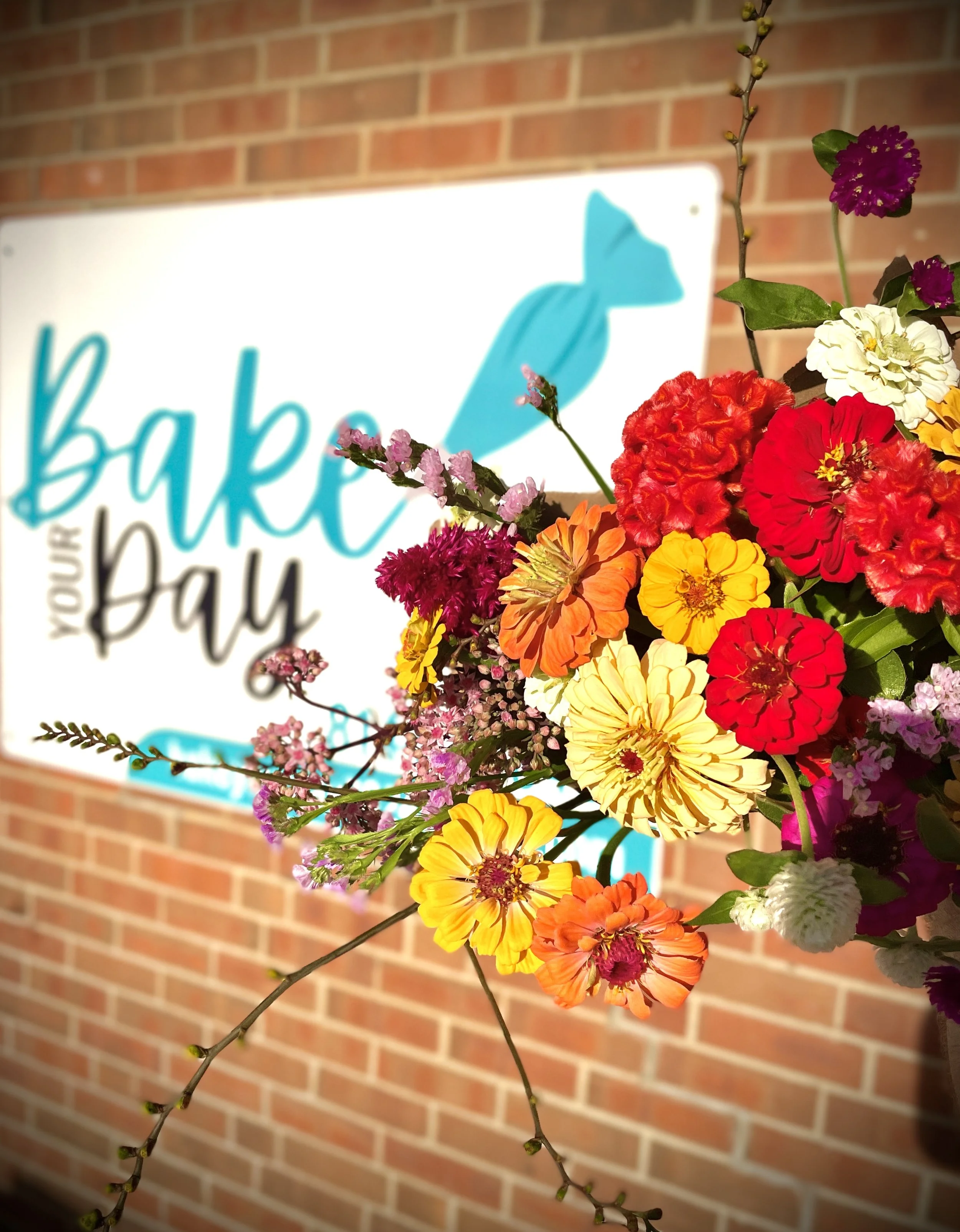 Colorful bouquet of flowers in front of a brick wall with a sign that says 'Bake Your Day' and features a blue bird logo.