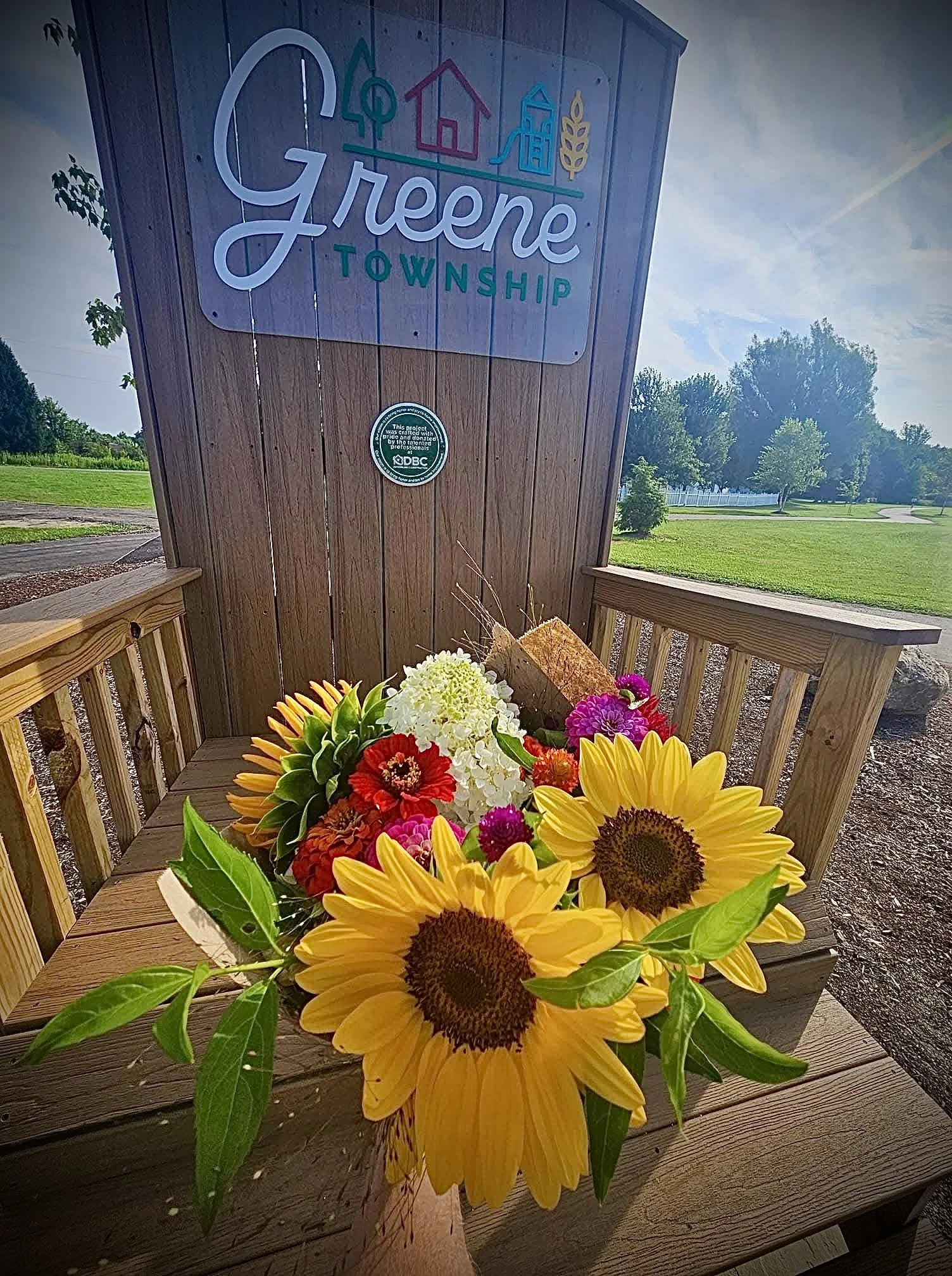 Colorful bouquet of sunflowers, red, white, pink, and orange flowers resting on a wooden bench outside in a park, with a sign for Greene Township in the background.