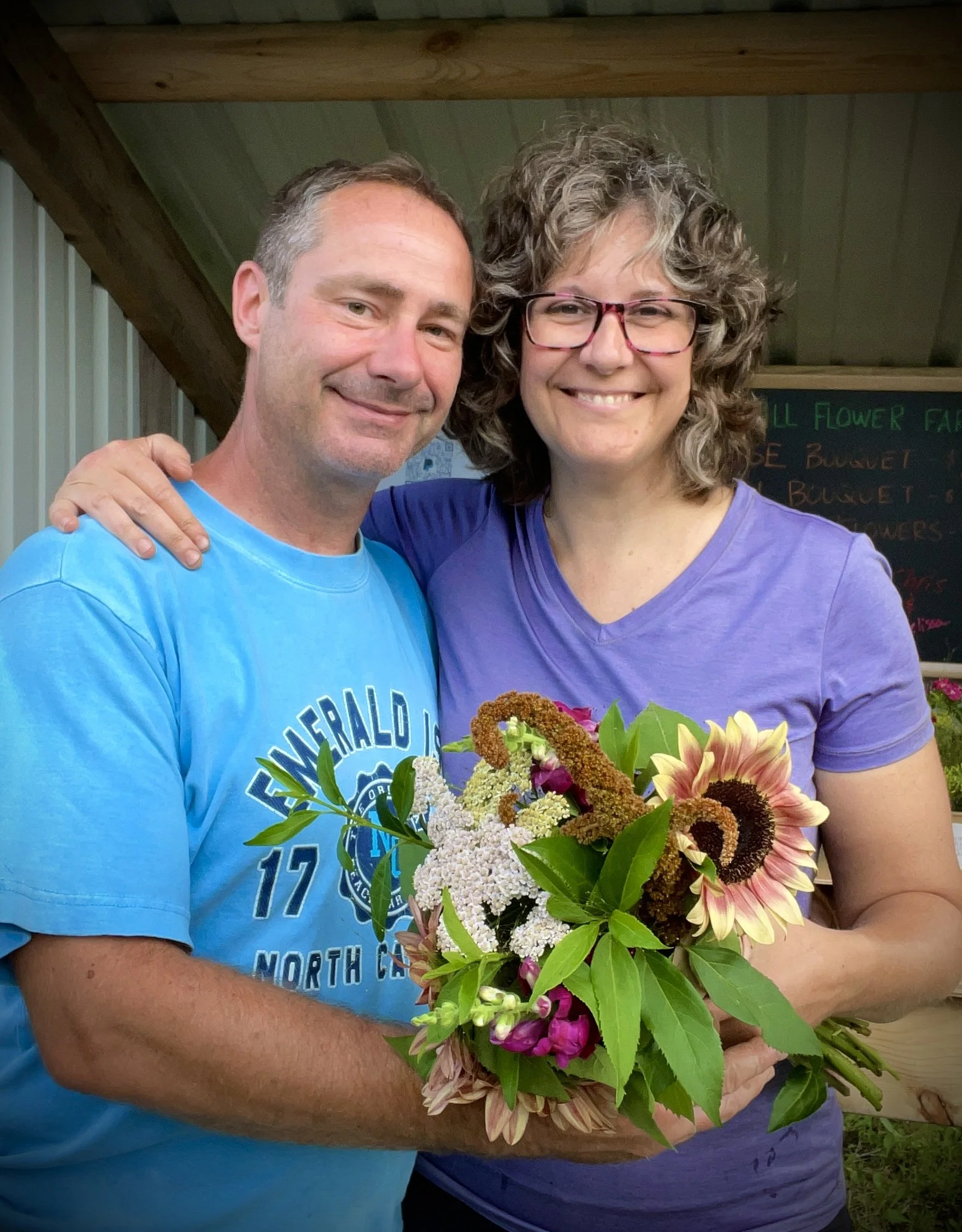 A man and woman smiling, holding a bouquet of flowers, standing indoors with a wooden ceiling and a chalkboard in the background.