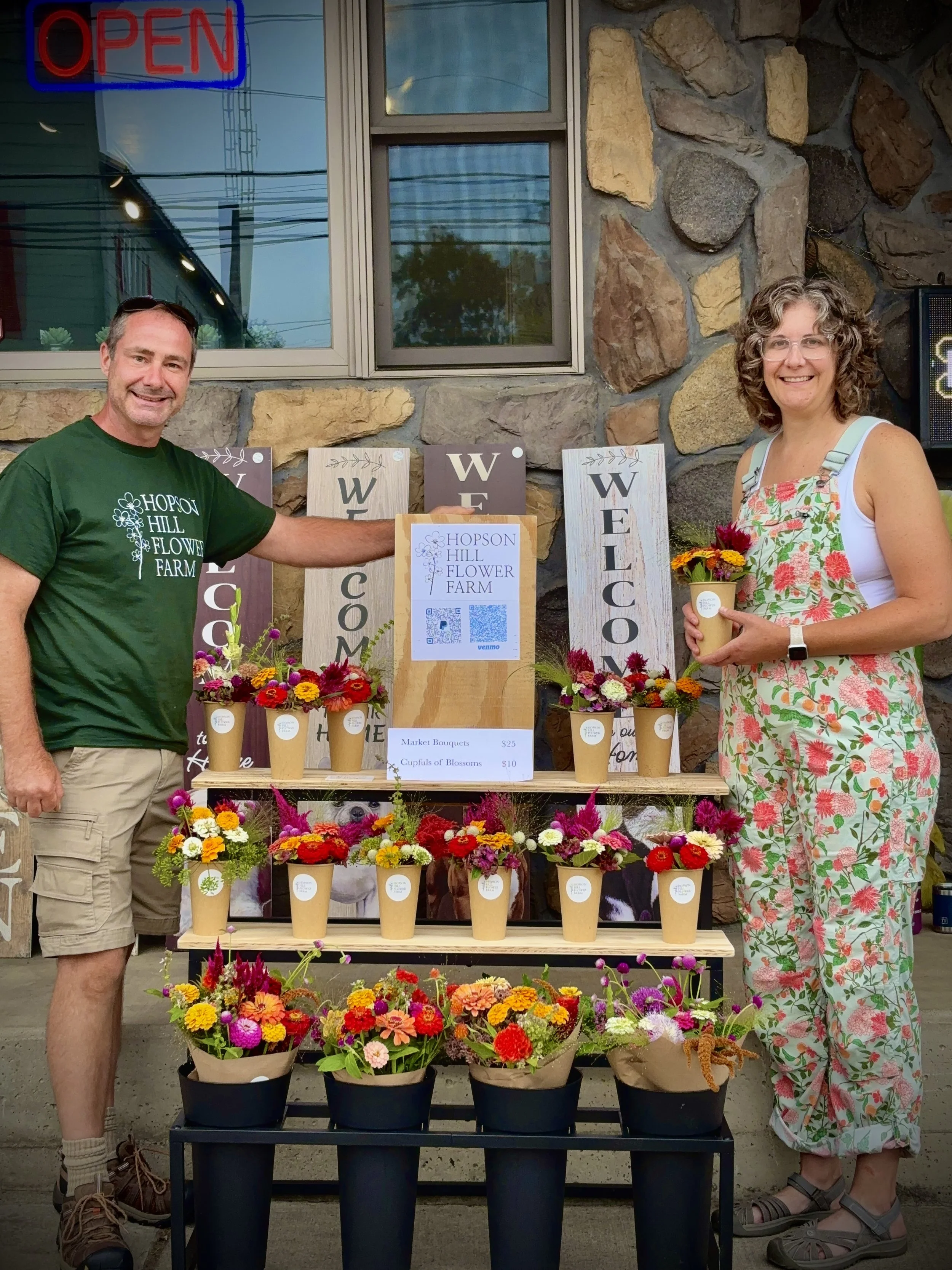 Two people standing in front of a flower display at Hopson Hill Flower Farm, with various colorful flower arrangements in pots, a welcome sign, and an 'Open' sign in the window behind them.