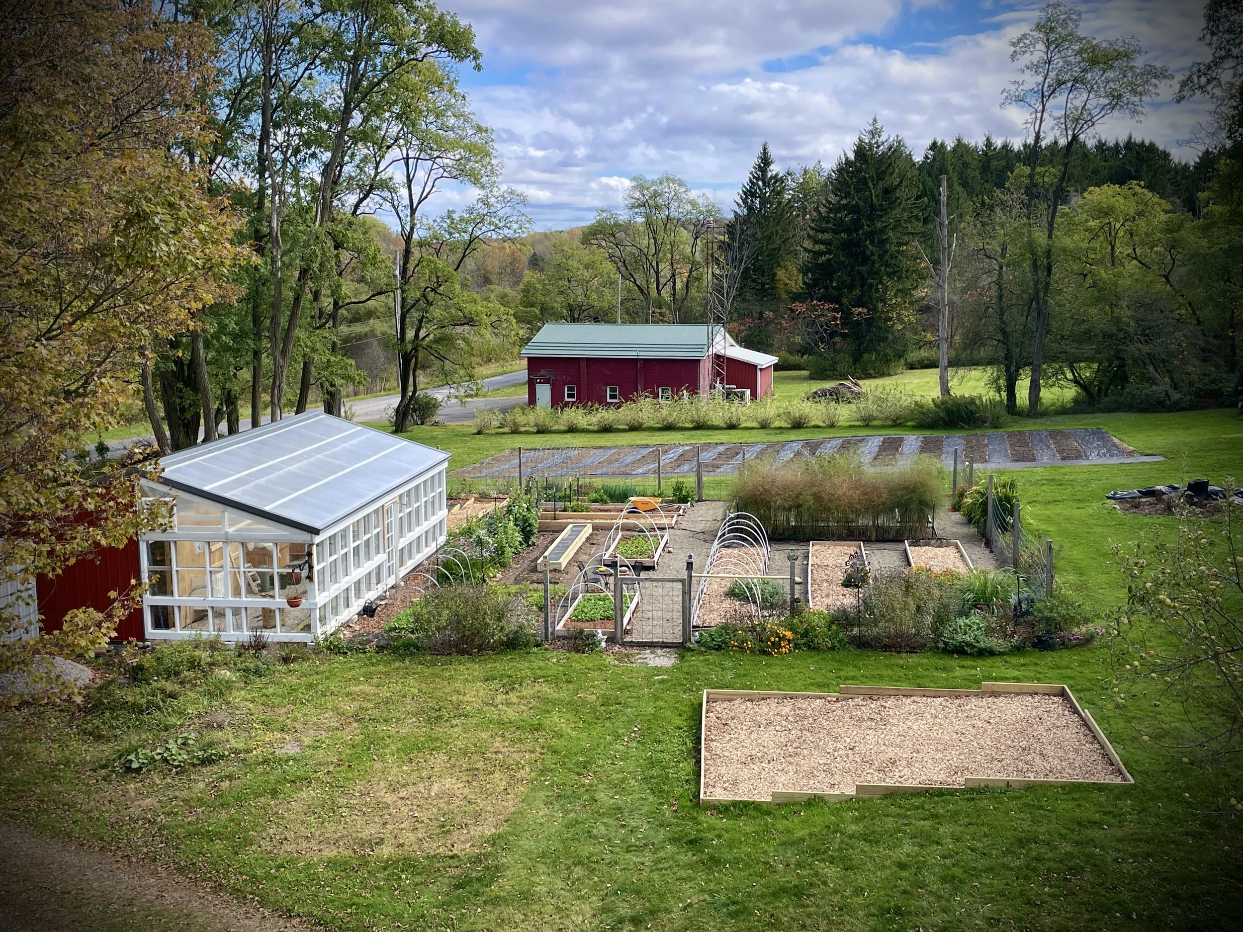 A garden with a greenhouse, multiple vegetable beds, and a red barn in the background amid a lush, green landscape with trees and a cloudy sky.
