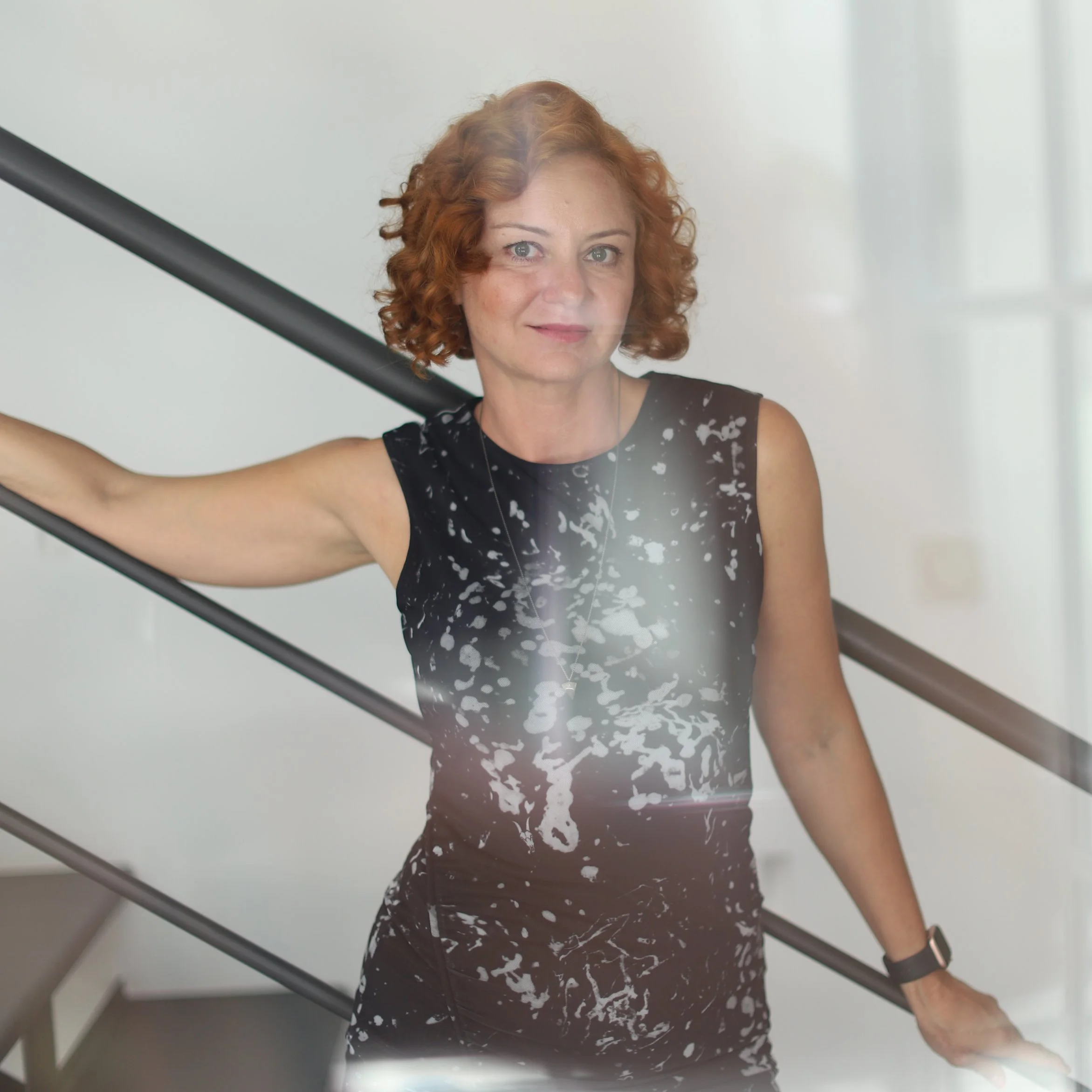 A woman with curly red hair and light skin, wearing a black sleeveless top with a white floral pattern, is holding a black rod or stick with her right hand and looking directly at the camera.
