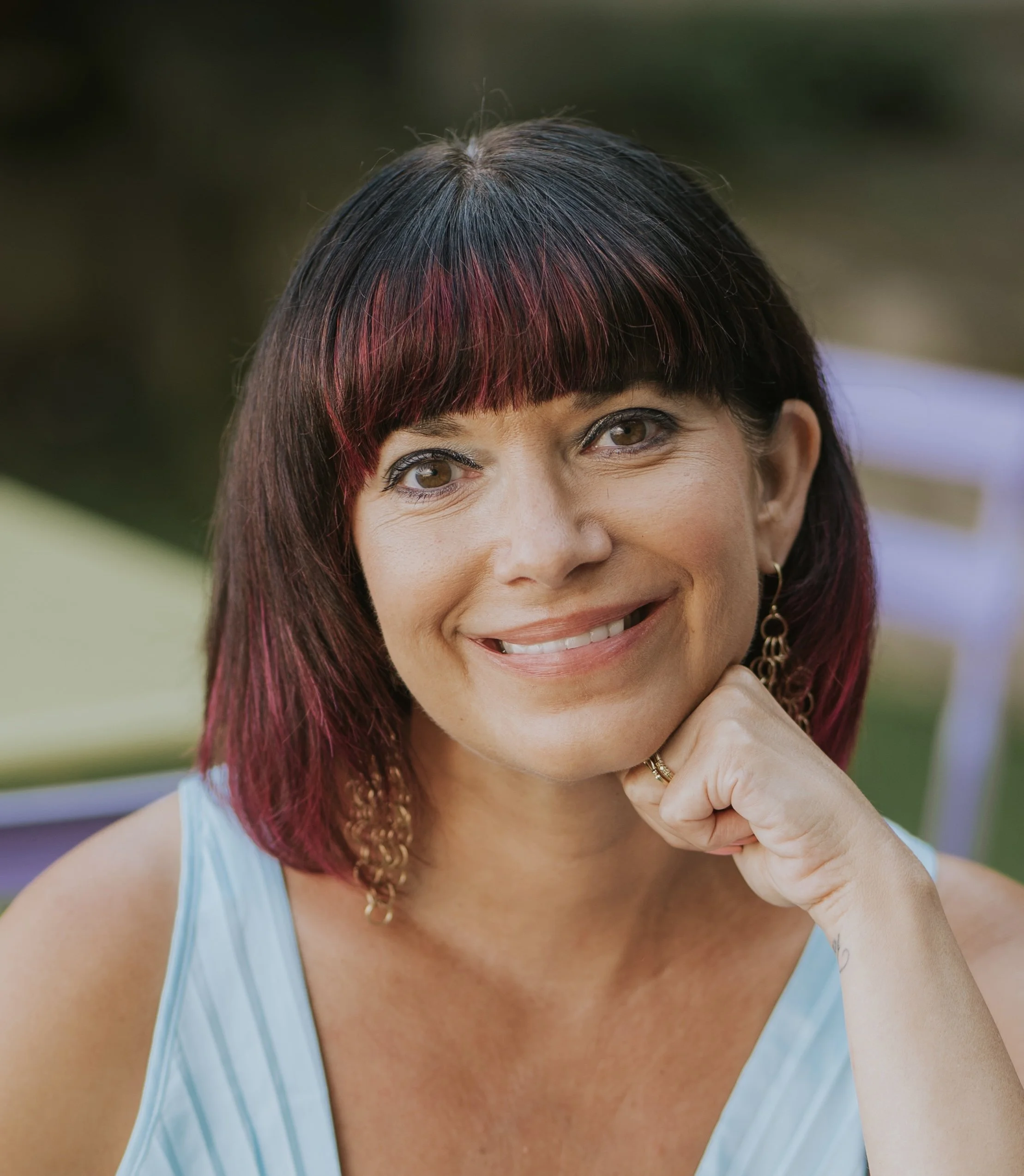A woman with short dark hair with red highlights, smiling and resting her chin on her hand, outdoors with blurred greenery in the background.