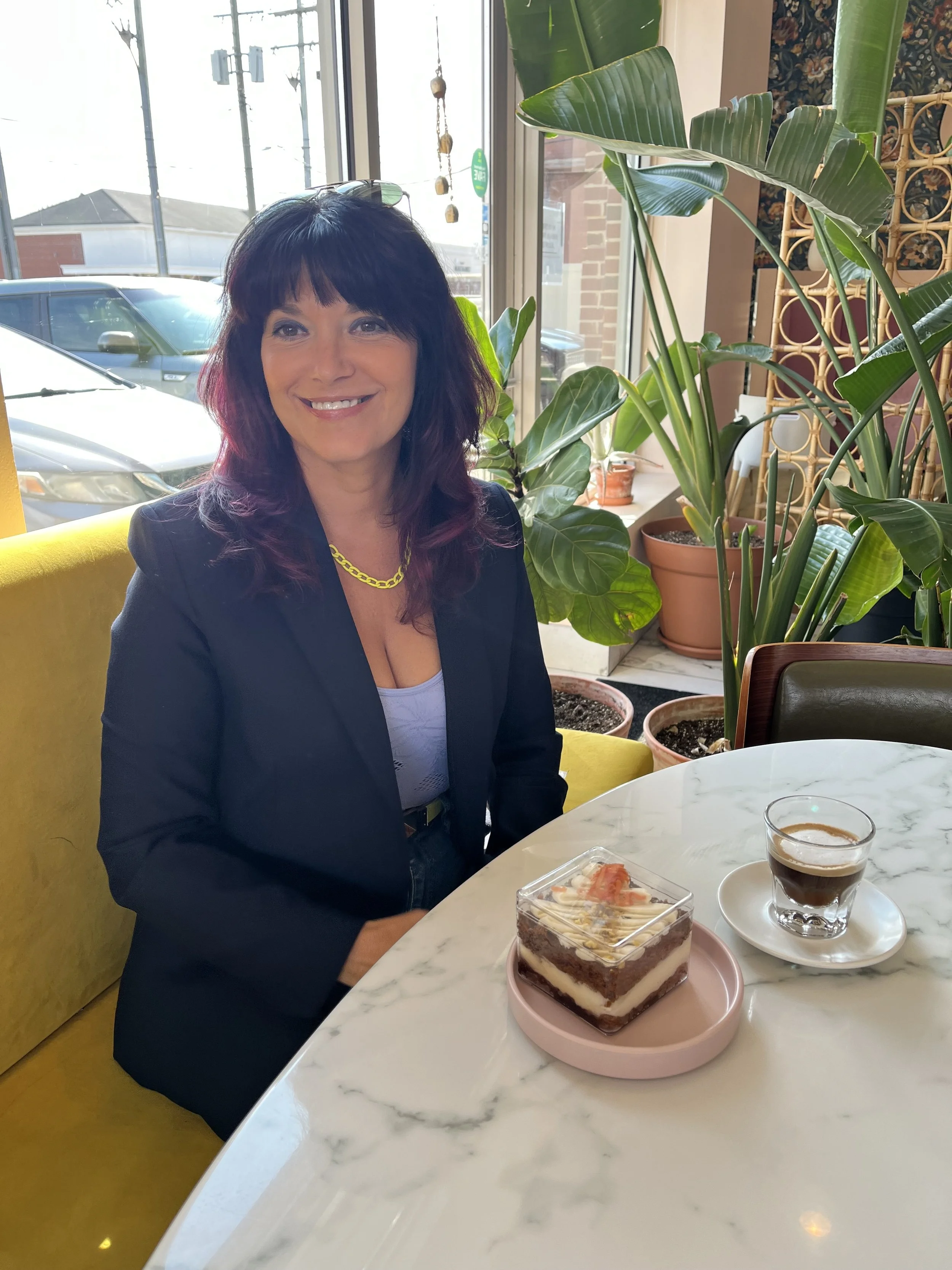 A woman with dark, purple-tinted hair smiling and sitting at a table in a cafe with a slice of layered cake and an espresso. She is wearing a black blazer, a light-colored top, a yellow chain necklace, and is seated next to a window with large green plants in the background.
