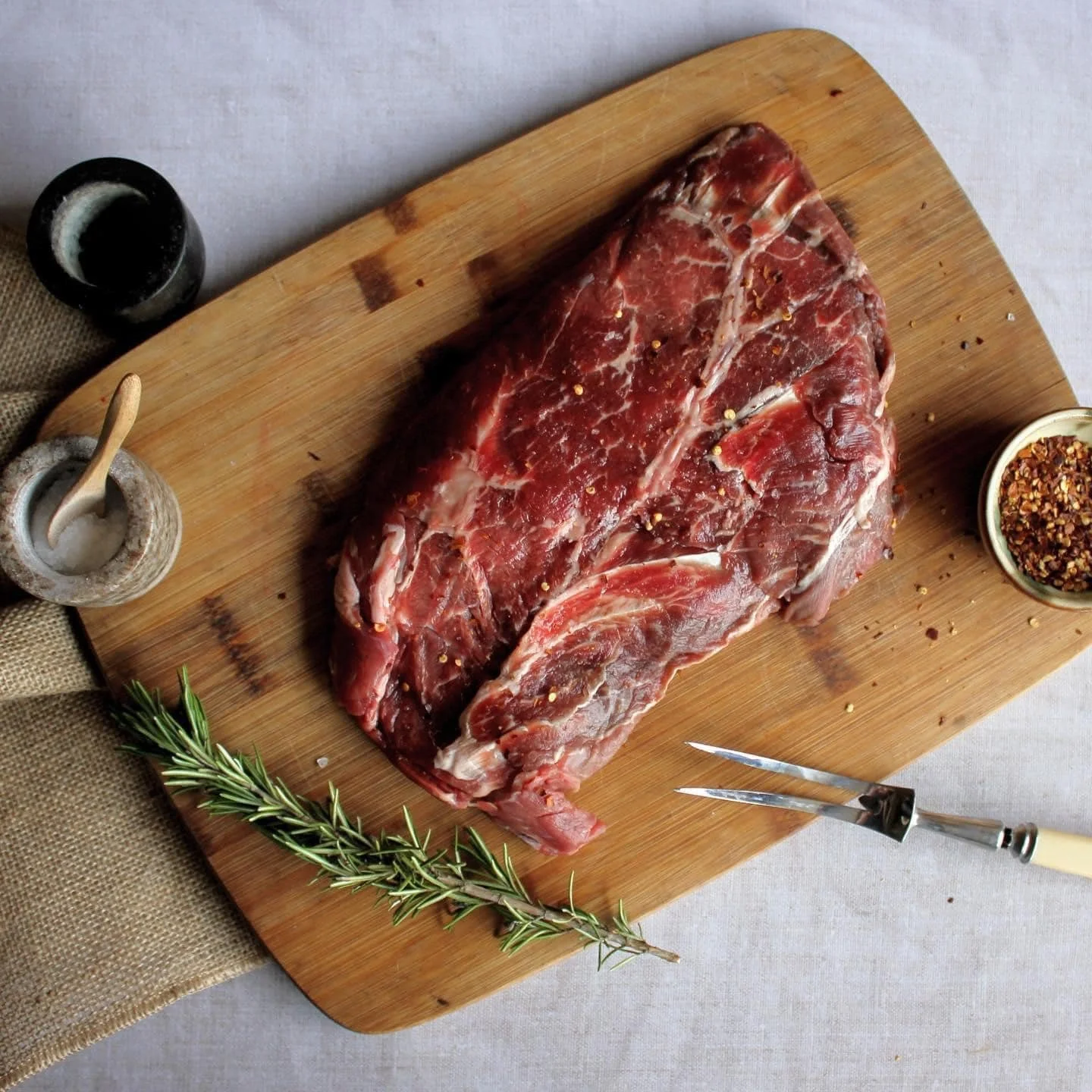 Raw beef steak with seasoning on a wooden cutting board, garnished with rosemary and accompanied by salt, pepper, and cooking utensils.