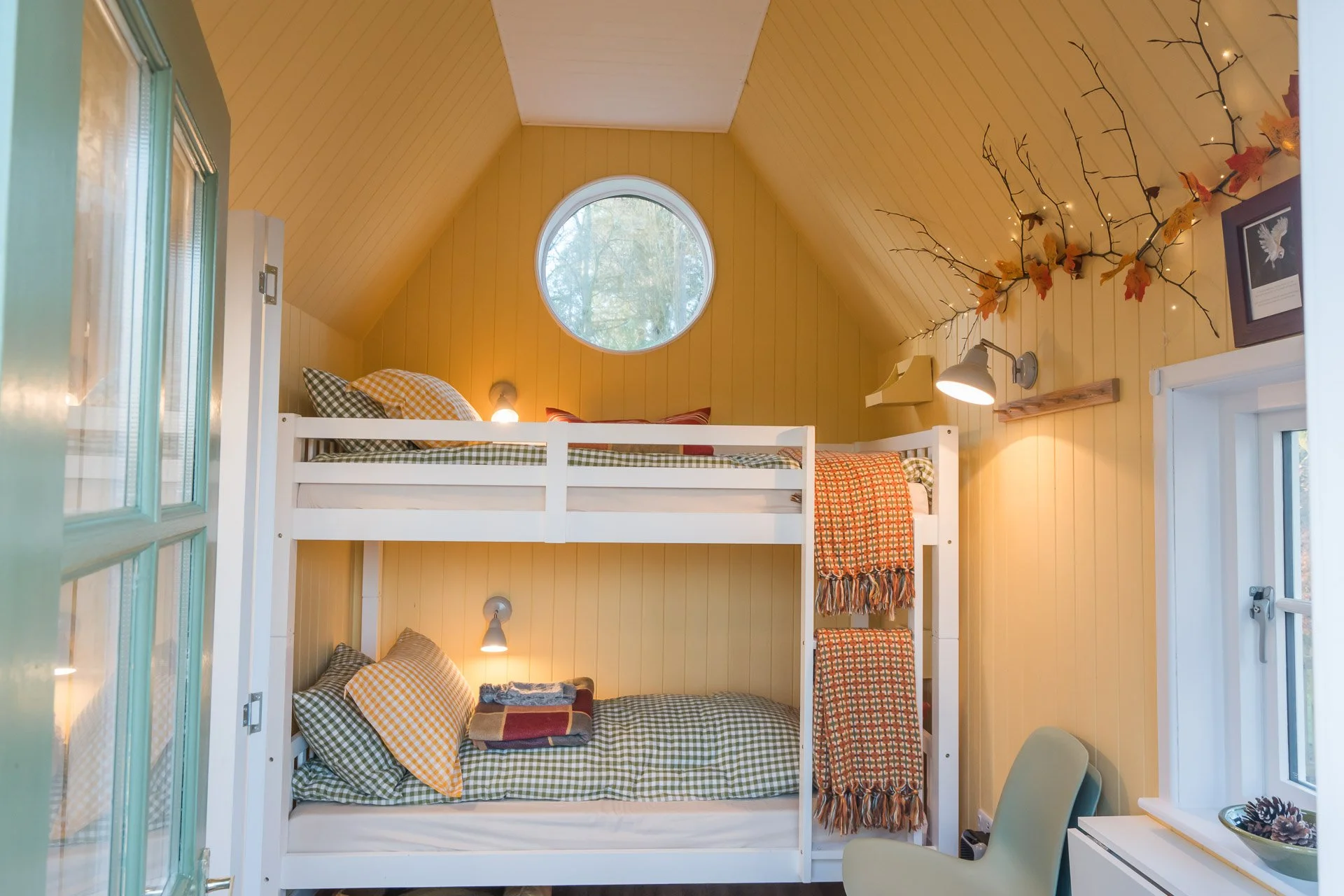 Cozy upstairs bedroom with yellow wooden walls, a white bunk bed, checkered bedding, a circular window, and autumn-themed decorations.