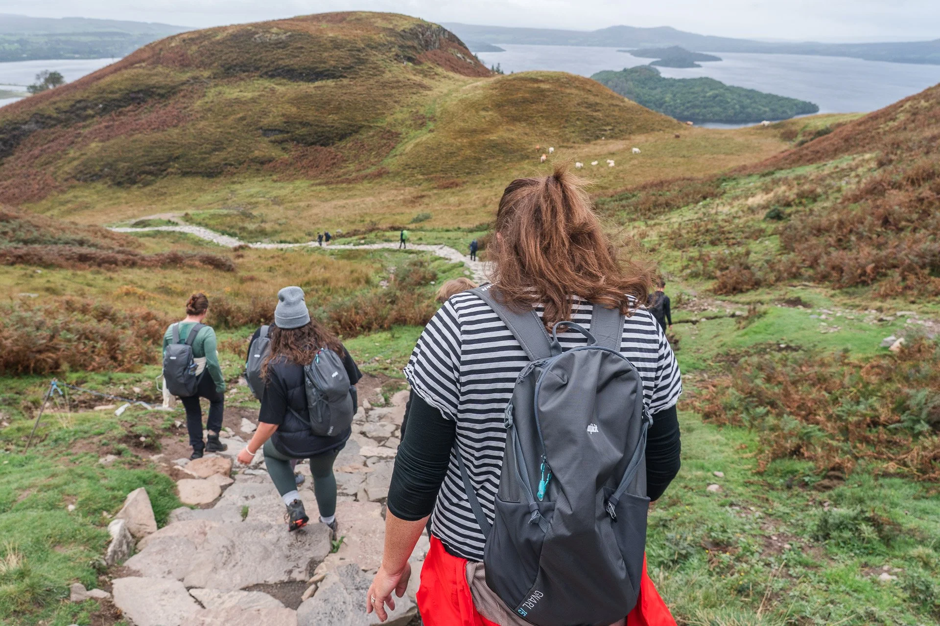 Group of hikers walking down a rocky trail through a scenic hilly landscape with green and brown vegetation, distant lake, and cloudy sky.
