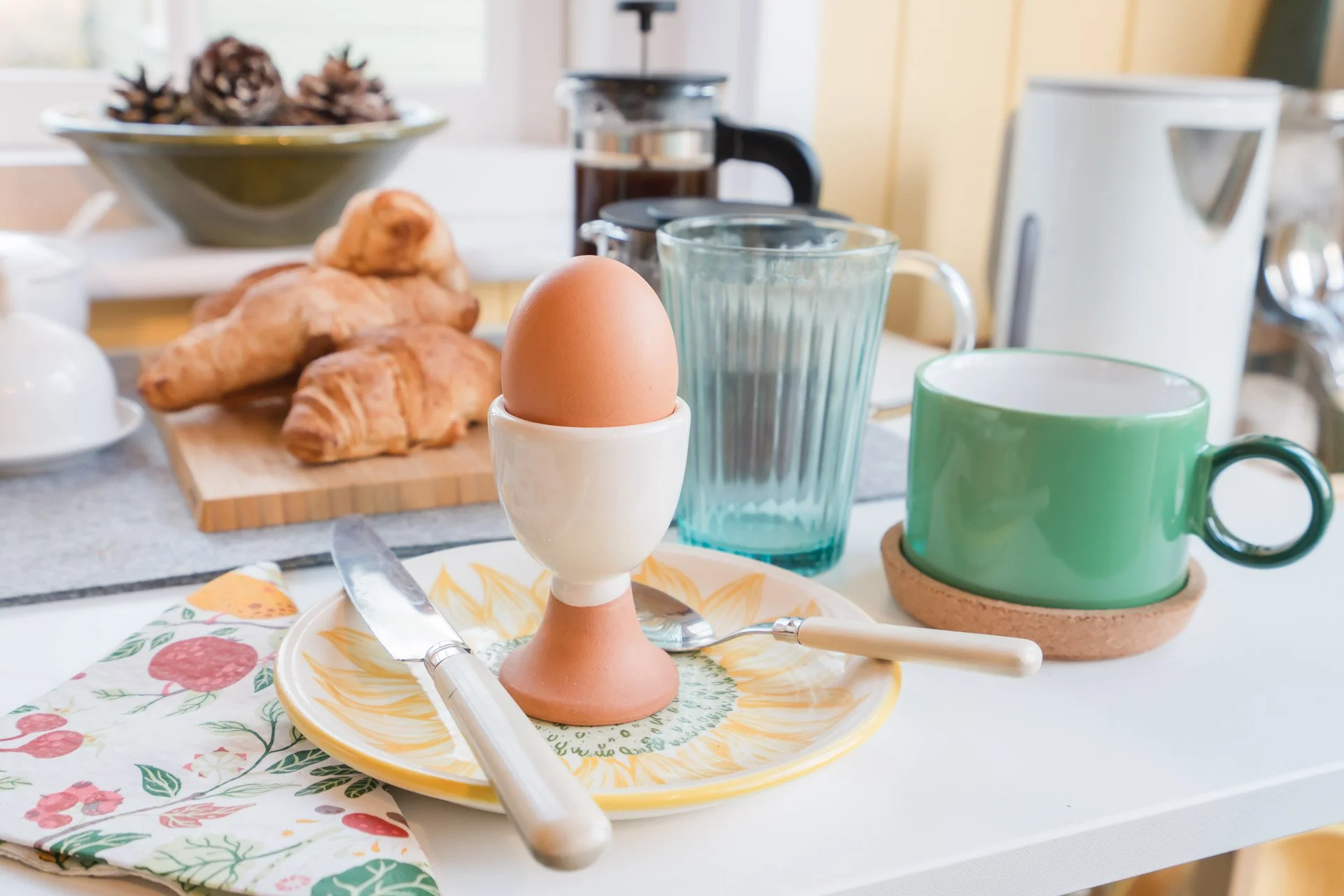 Breakfast setup with a soft-boiled egg in an egg cup, a plate with a knife and spoon, a colorful napkin, croissants, a green mug on a cork coaster, an empty glass, a pitcher, a French press, and a coffee maker on a kitchen counter.