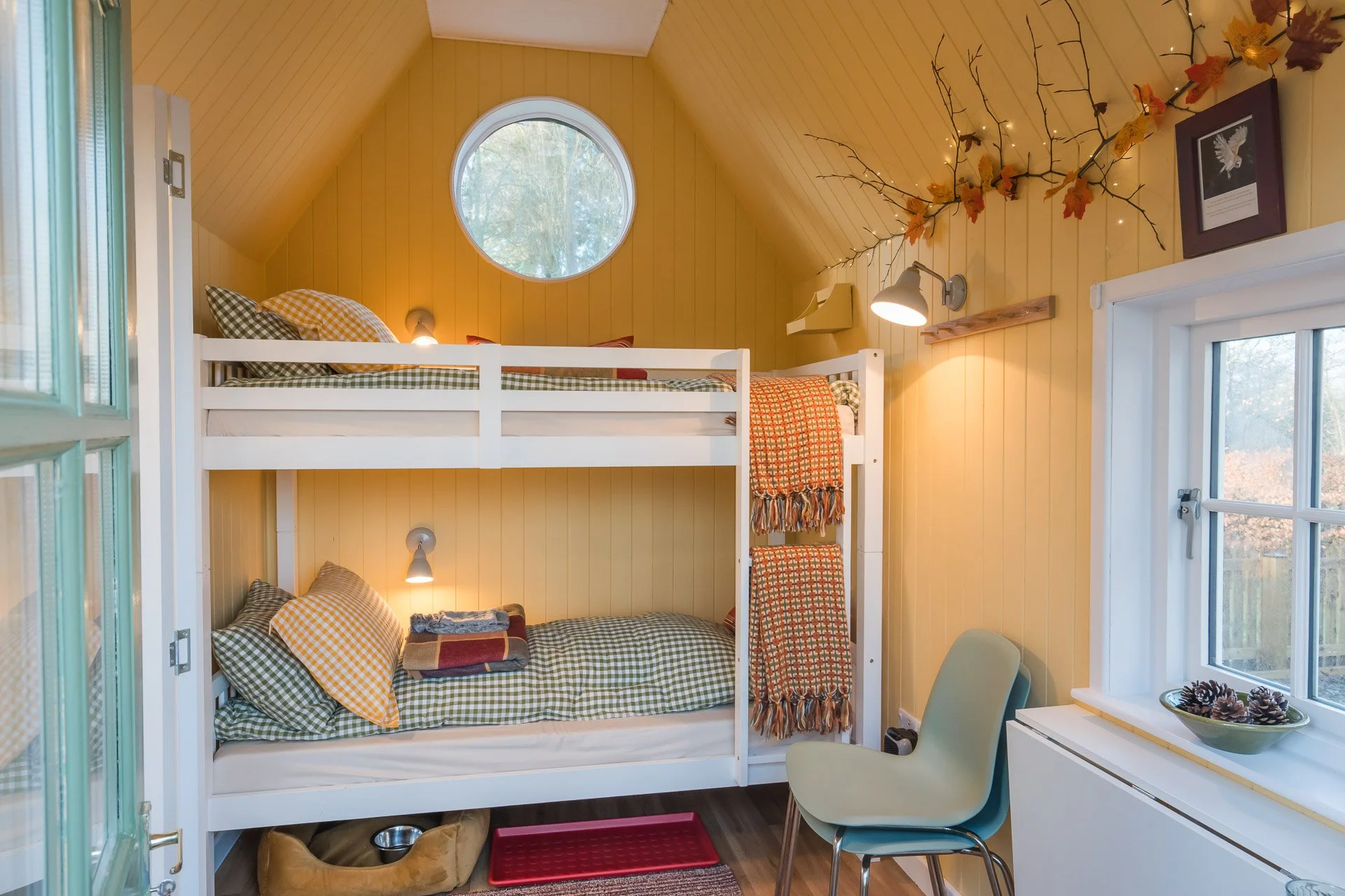 Interior of a cozy bedroom with yellow walls, a white bunk bed with checkered and patterned bedding, large round window, decorative branch with autumn leaves and string lights, framed artwork, chair, and window with a bowl of pinecones.