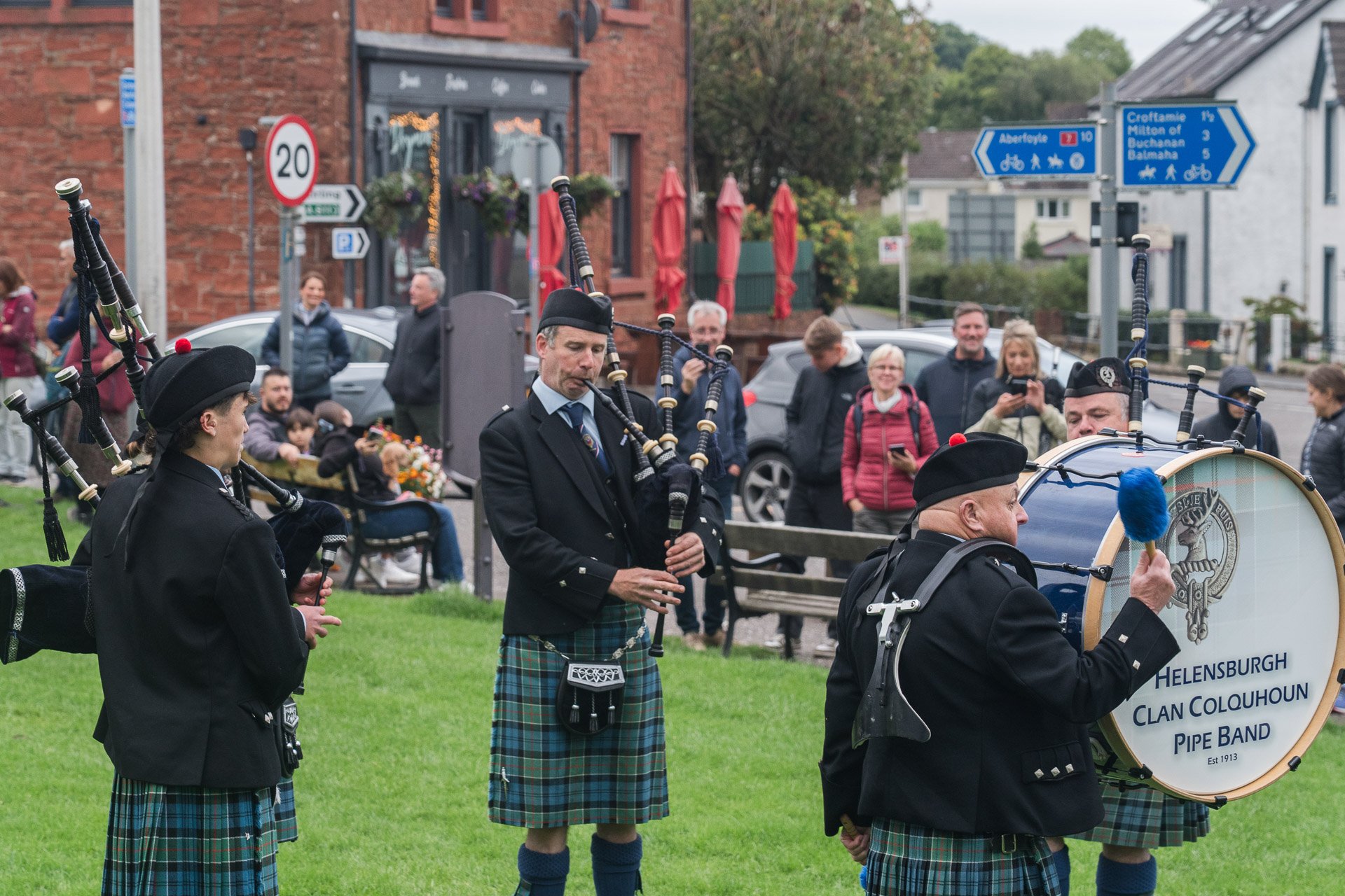 A group of musicians in traditional Scottish attire playing pipes and drums outdoors, with onlookers watching in the background.