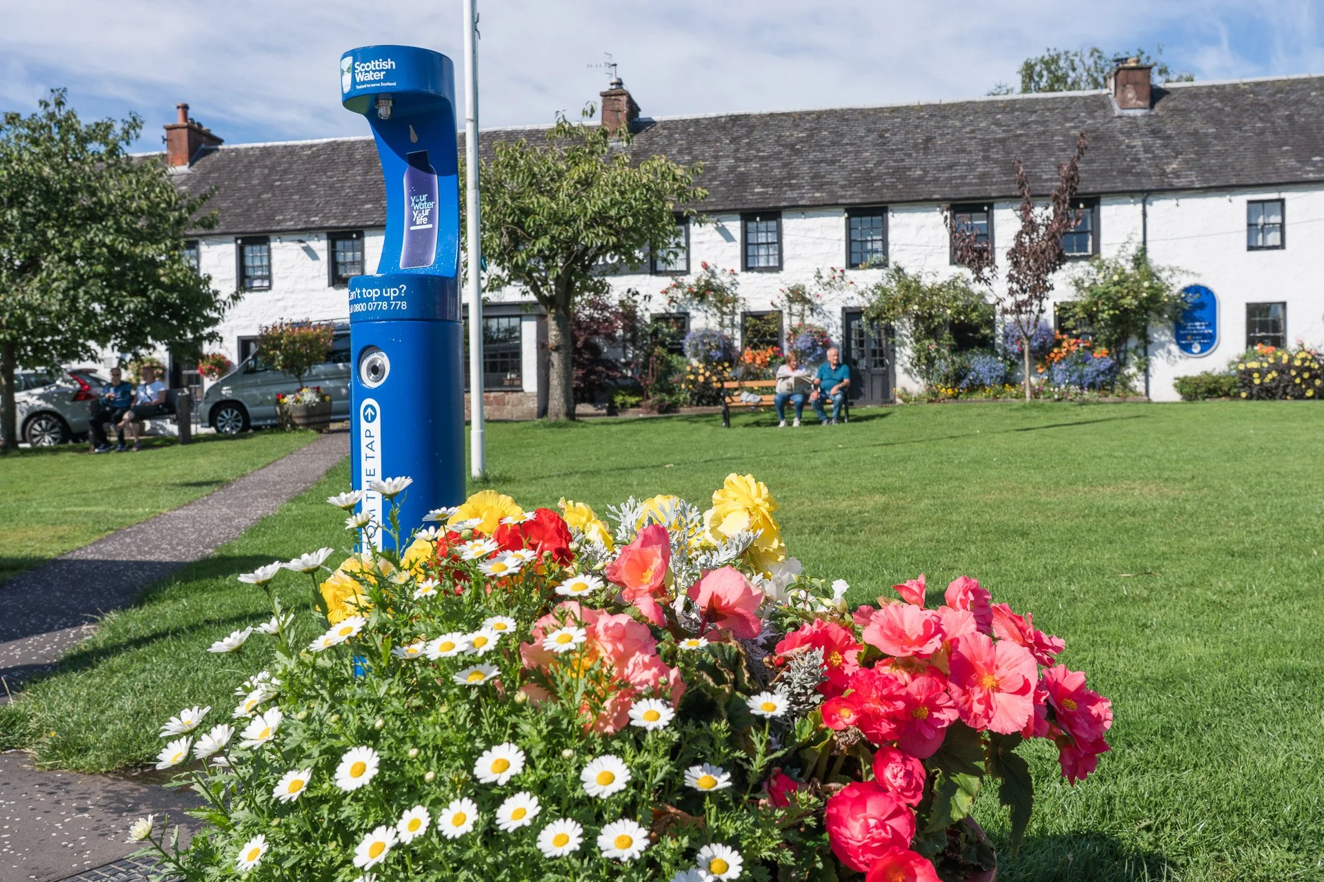 A blue Scottish Water vending station in a park with colorful flowers in the foreground and a white building with people sitting on benches in the background.