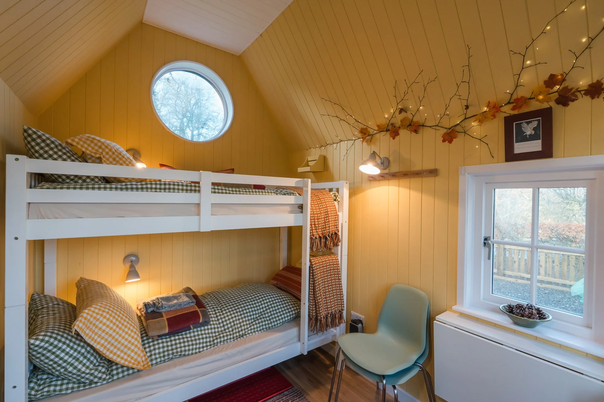 A cozy bedroom with upper and lower bunk beds with checkered green and yellow bedding, yellow wooden walls, a small round window, a chair, and window with a decorative bowl of pinecones.