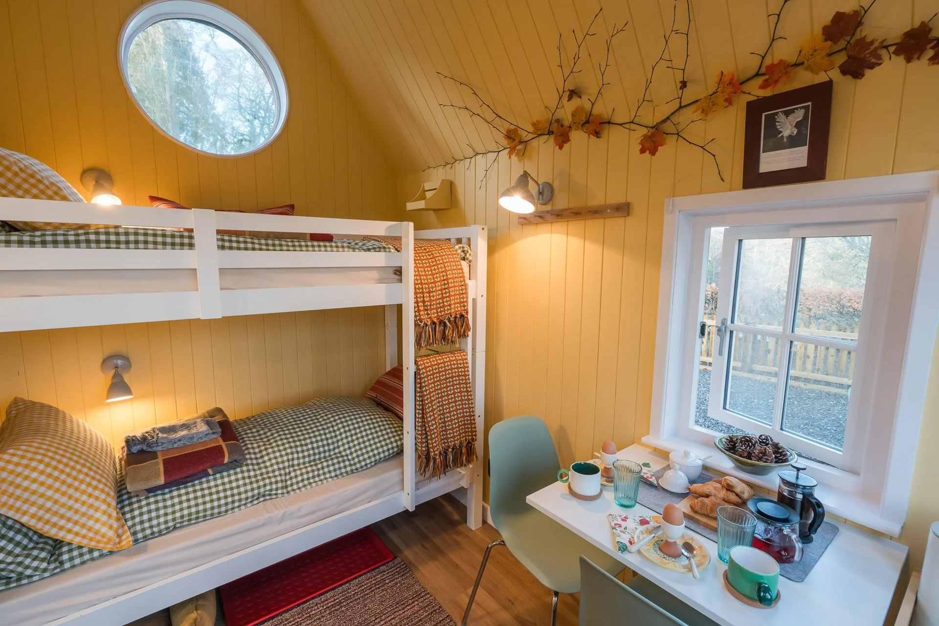 A cozy bedroom with wooden yellow paneled walls, featuring a bunk bed with checkered green and white bedding, a small white table with breakfast items, and windows showing trees outside.