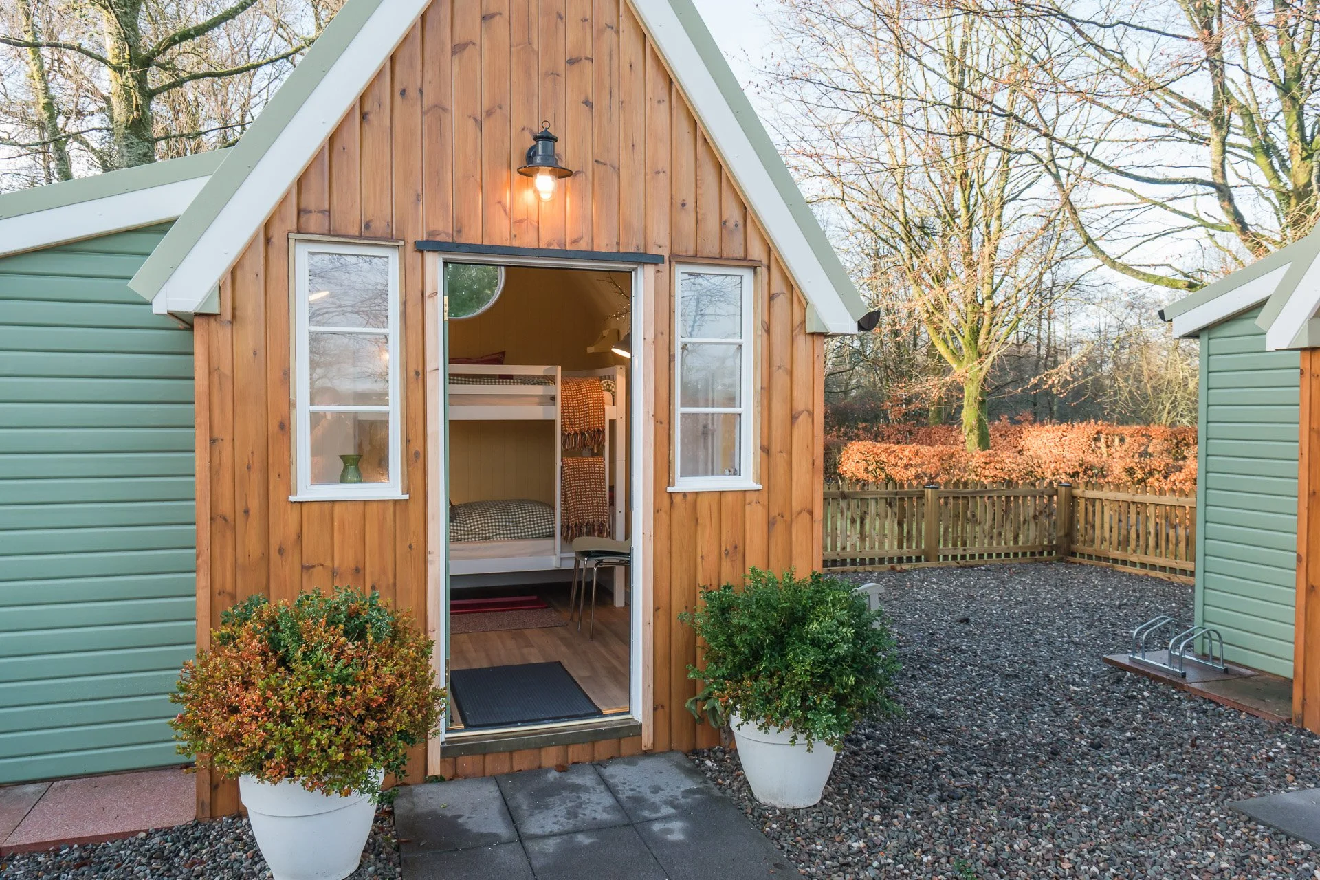 A small cabin with wooden siding and white-trimmed windows, open door revealing bunk beds inside, surrounded by potted bushes and a gravel yard, with a wooden fence and leafless trees in the background.