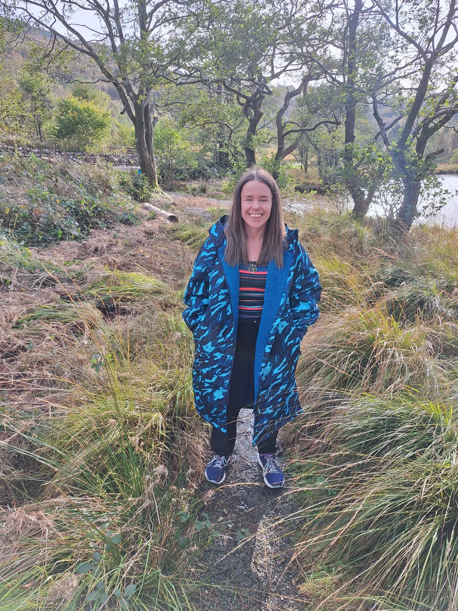A young woman stands on a narrow trail in a natural outdoor setting with trees and grassy plants around her, smiling at the camera.