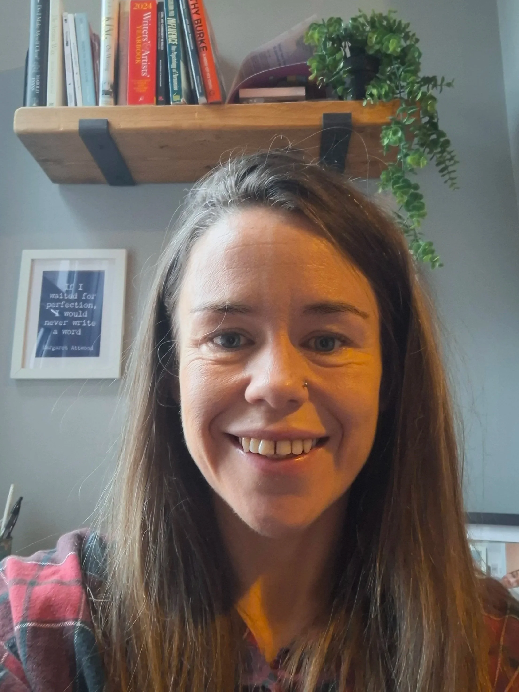 A woman with long brown hair smiling in front of a gray wall with a framed quote, a wooden shelf with books, and a green potted plant.