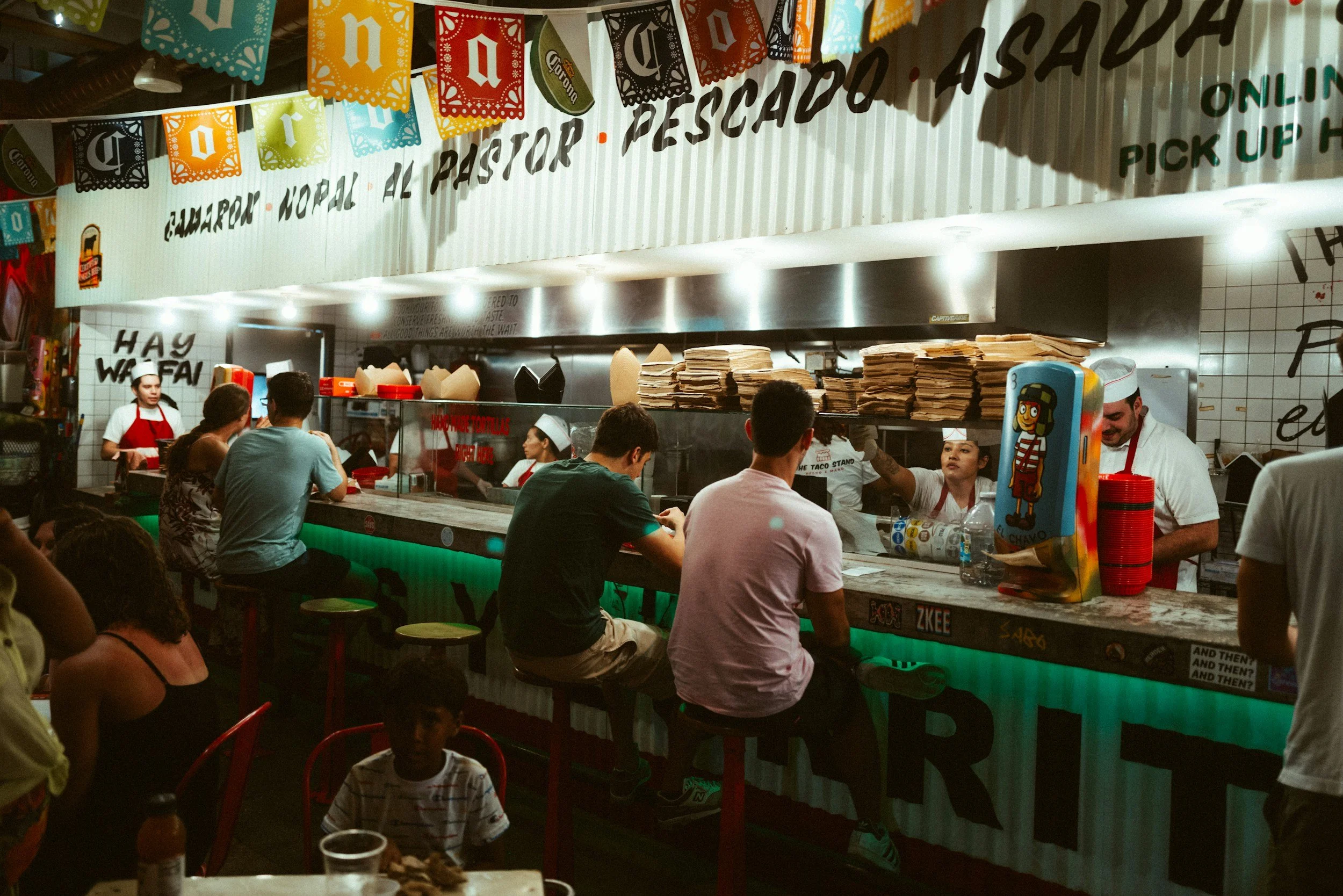 People sitting and ordering food at a colorful taco restaurant with a counter and staff preparing orders.