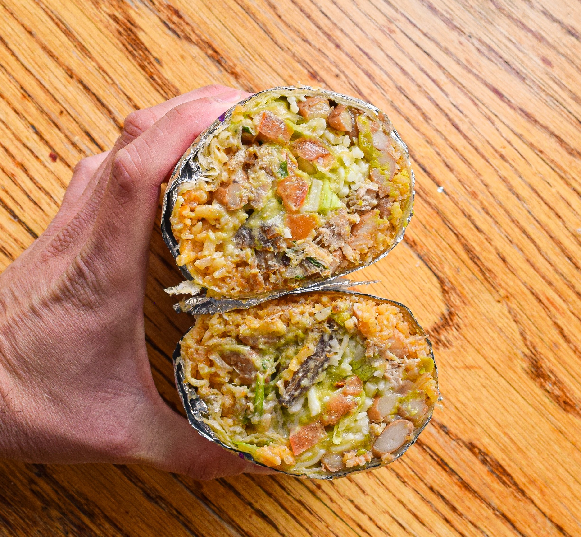 A hand holding two halves of a burrito with rice, vegetables, and meat, on a wooden surface.