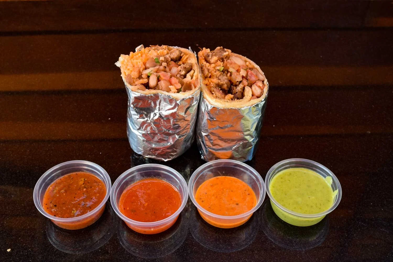Cut carne asada burrito wrapped in foil with four salsa cups—fire-roasted, roja, habanero, and verde—arranged in front.