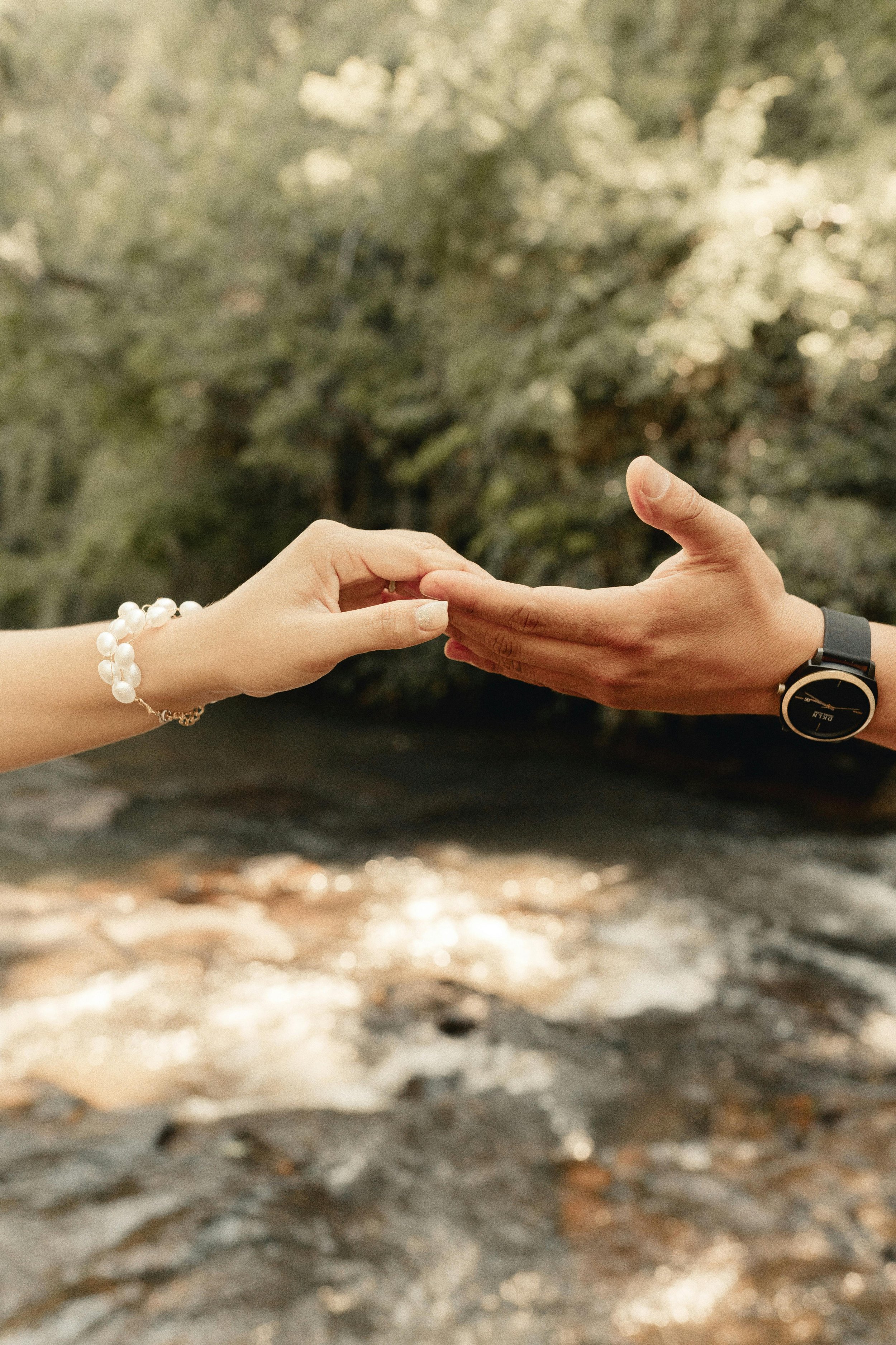Two people reaching out to touch each other's hands outdoors near a river surrounded by trees.