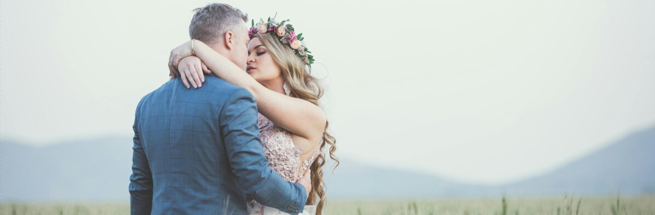 A couple embracing outdoors, with the woman wearing a flower crown and a lace dress, and the man in a blue suit.