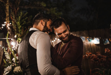 Two men hugging at night during a celebration or wedding, with festive lights and flowers in the background.