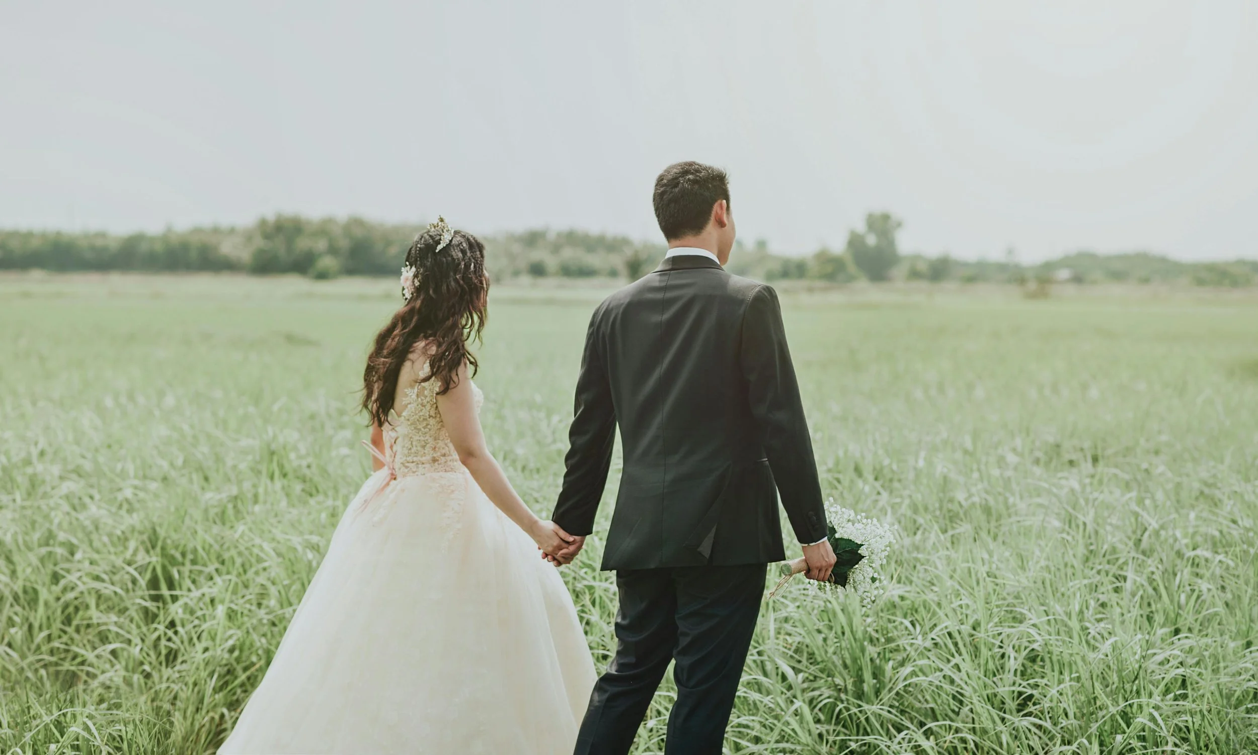 A newlywed couple holding hands in a green field with the groom holding a bouquet of flowers, facing away from the camera under a bright sky.