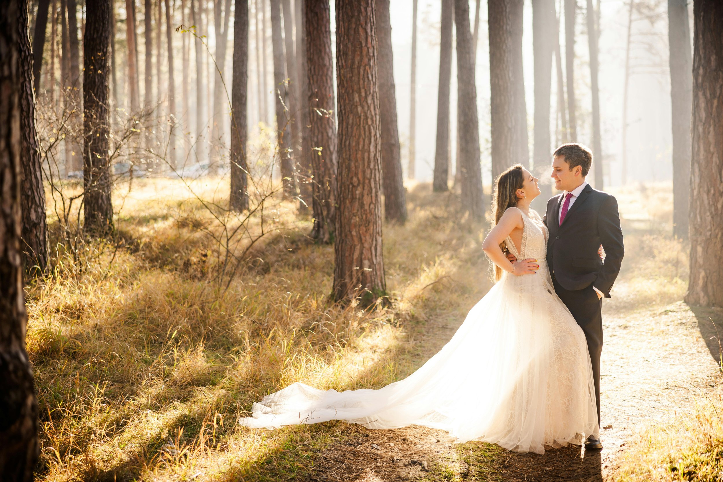 A bride and groom standing face-to-face on a forest path, smiling at each other, with sunlight filtering through tall trees.