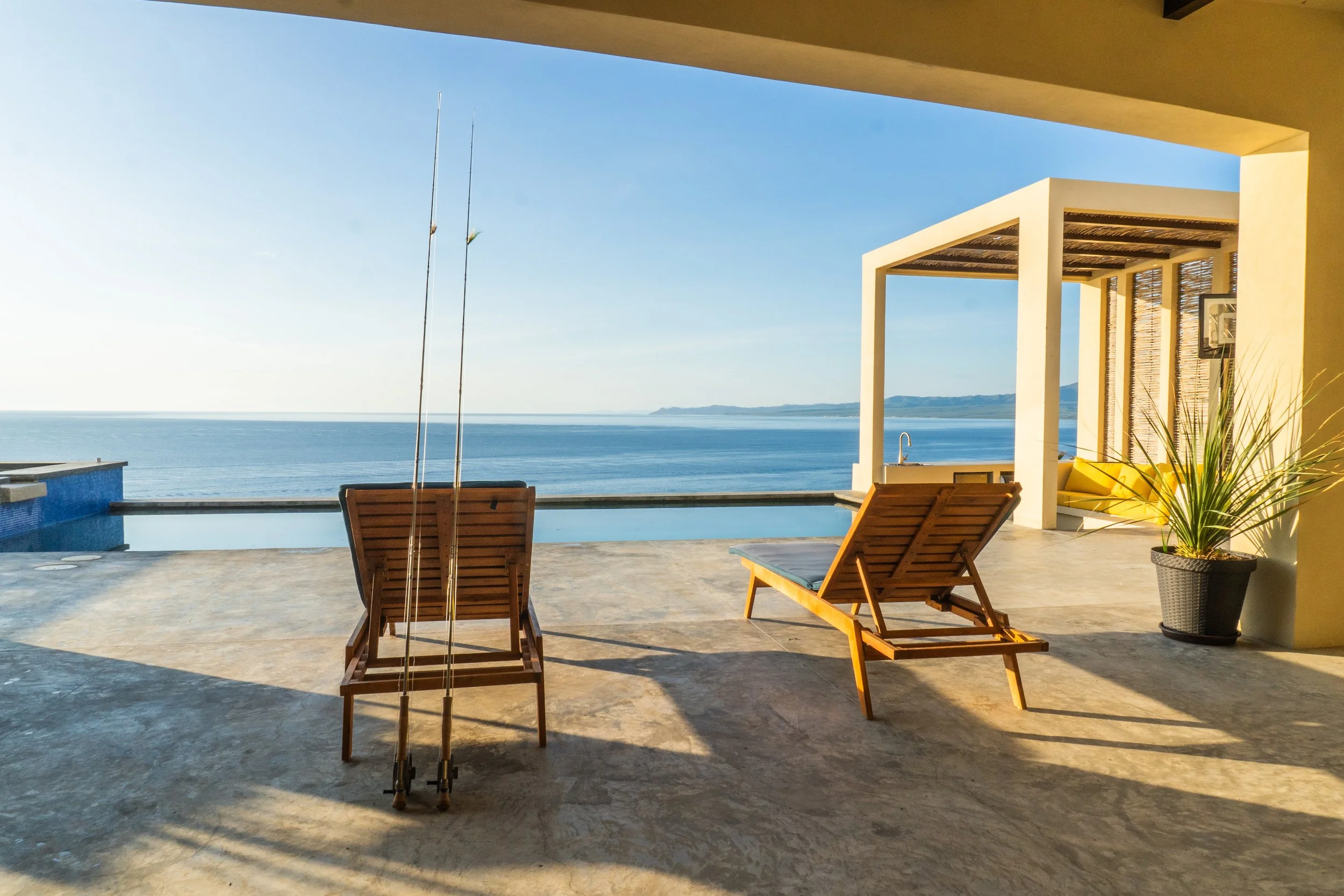 Balcony with two wooden lounge chairs facing the ocean under a covered patio, with fishing poles leaning against one chair, and a potted plant to the right, overlooking a blue ocean and distant hills.