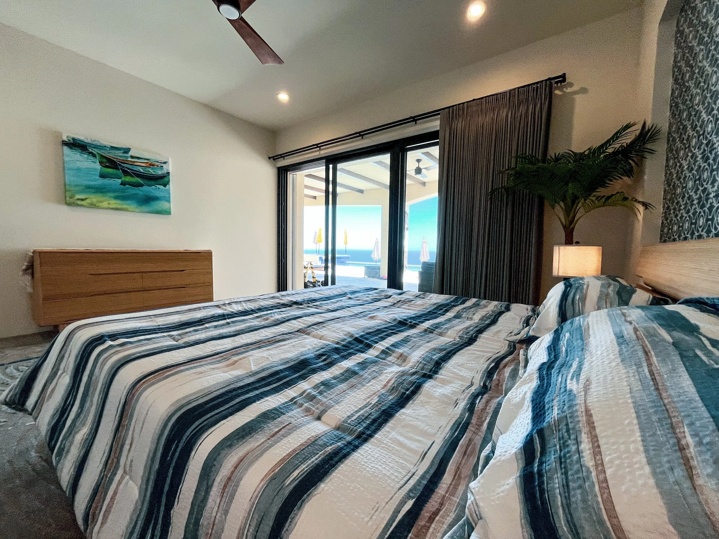 A bedroom with a large bed covered in a striped blue, beige, and white comforter. There are pillows on the bed. To the right, a nightstand with a lamp and a large potted plant. A sliding glass door opens to a patio with an ocean view, visible with beach umbrellas. A ceiling fan and recessed ceiling lights are also visible.