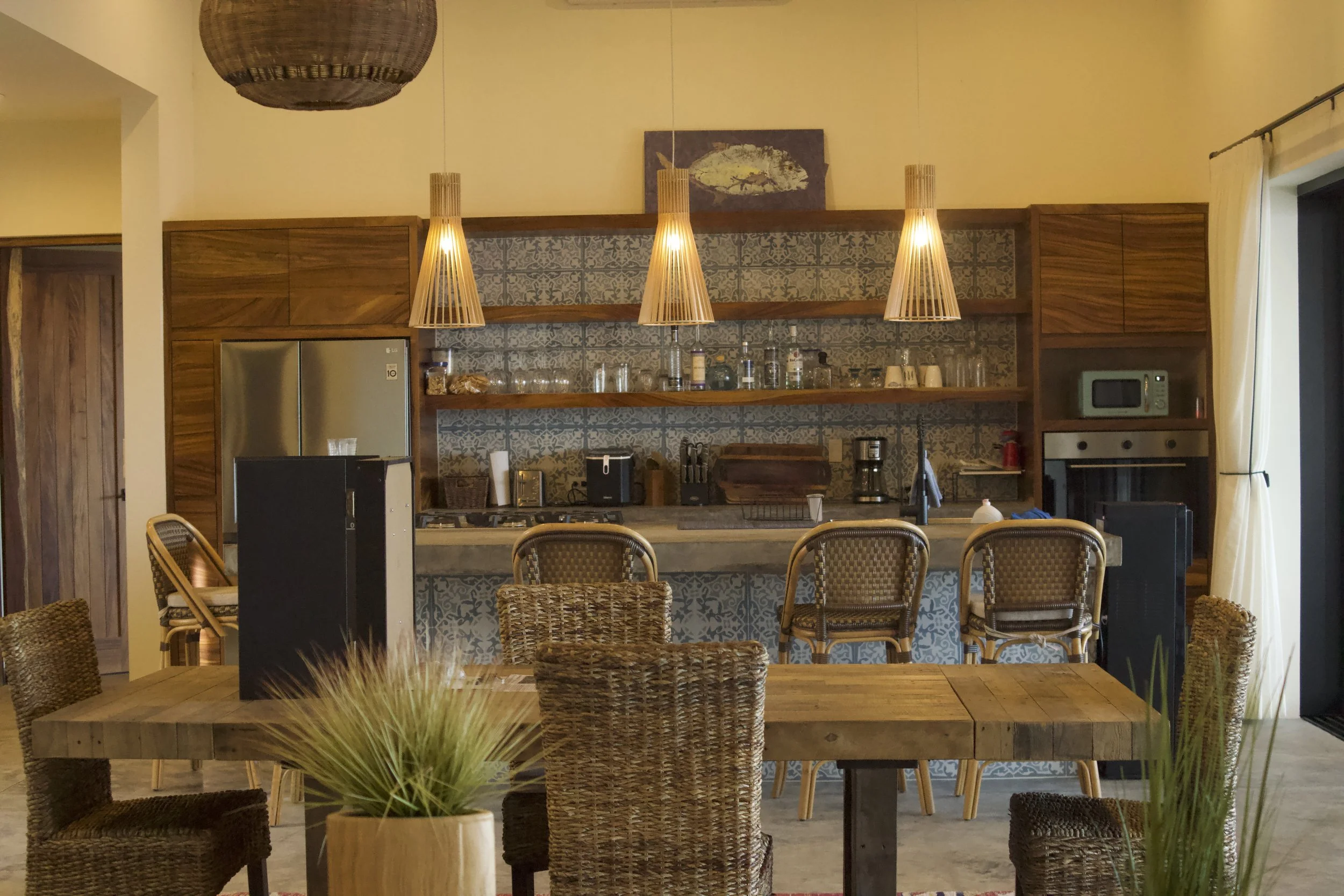 A cozy kitchen with wooden cabinets, patterned backsplash, open shelves with glassware, and a concrete countertop. Wooden pendant lights hang above the counter, and a dining table with wicker chairs is in the foreground, decorated with potted plants.