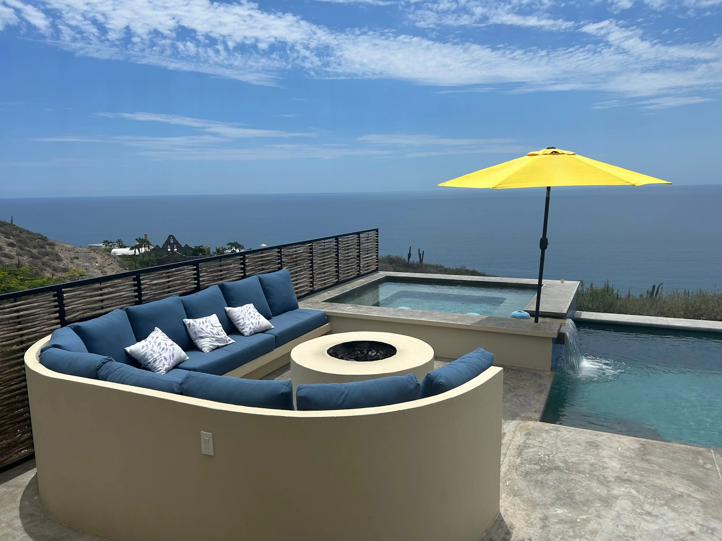 Outdoor patio with blue cushioned seating, a yellow umbrella over a hot tub, a nearby pool with a waterfall feature, and an ocean view in the background.