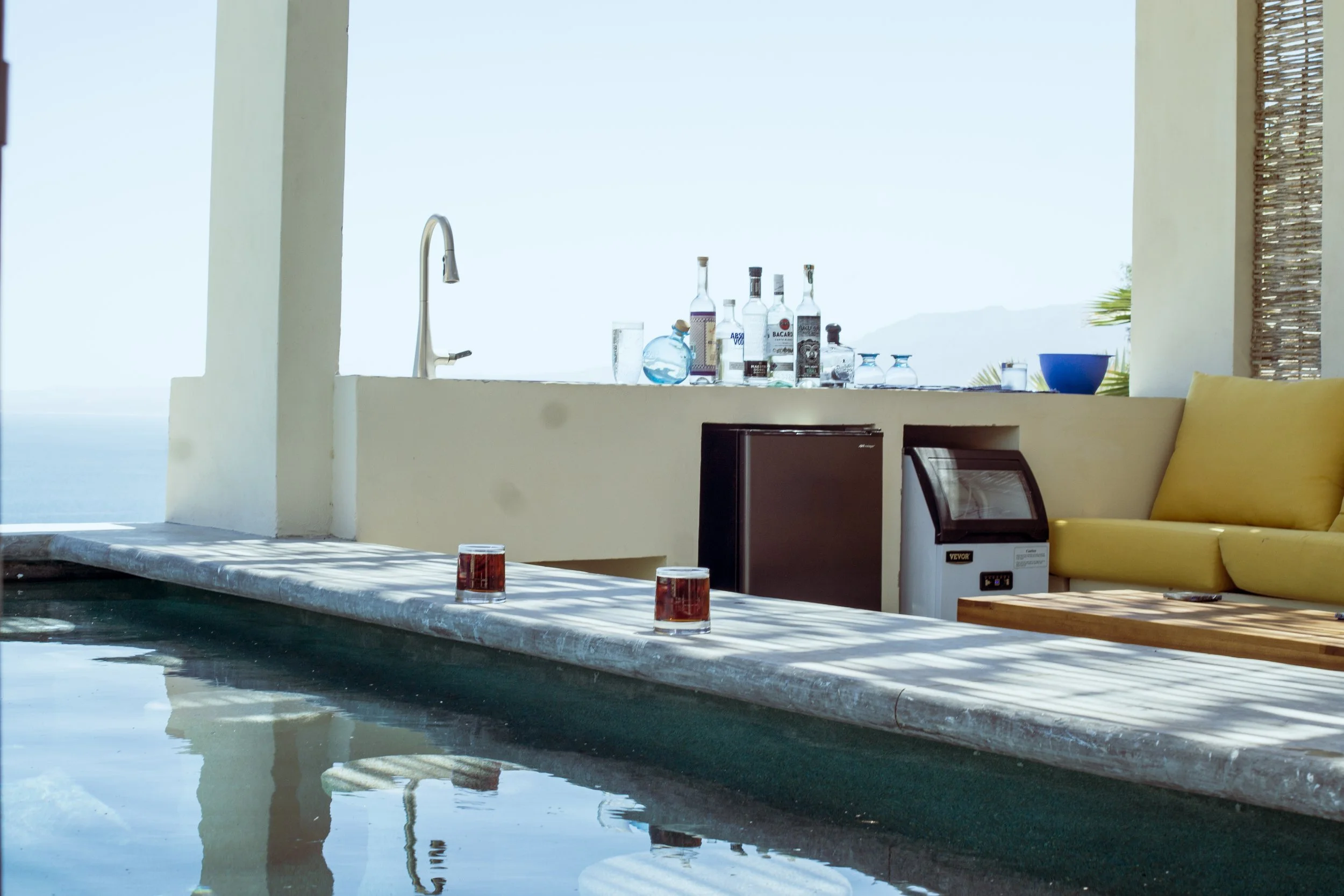 A poolside bar area with bottles of liquor, glassware, a sink, and seating with a yellow cushion, overlooking the ocean on a clear day.