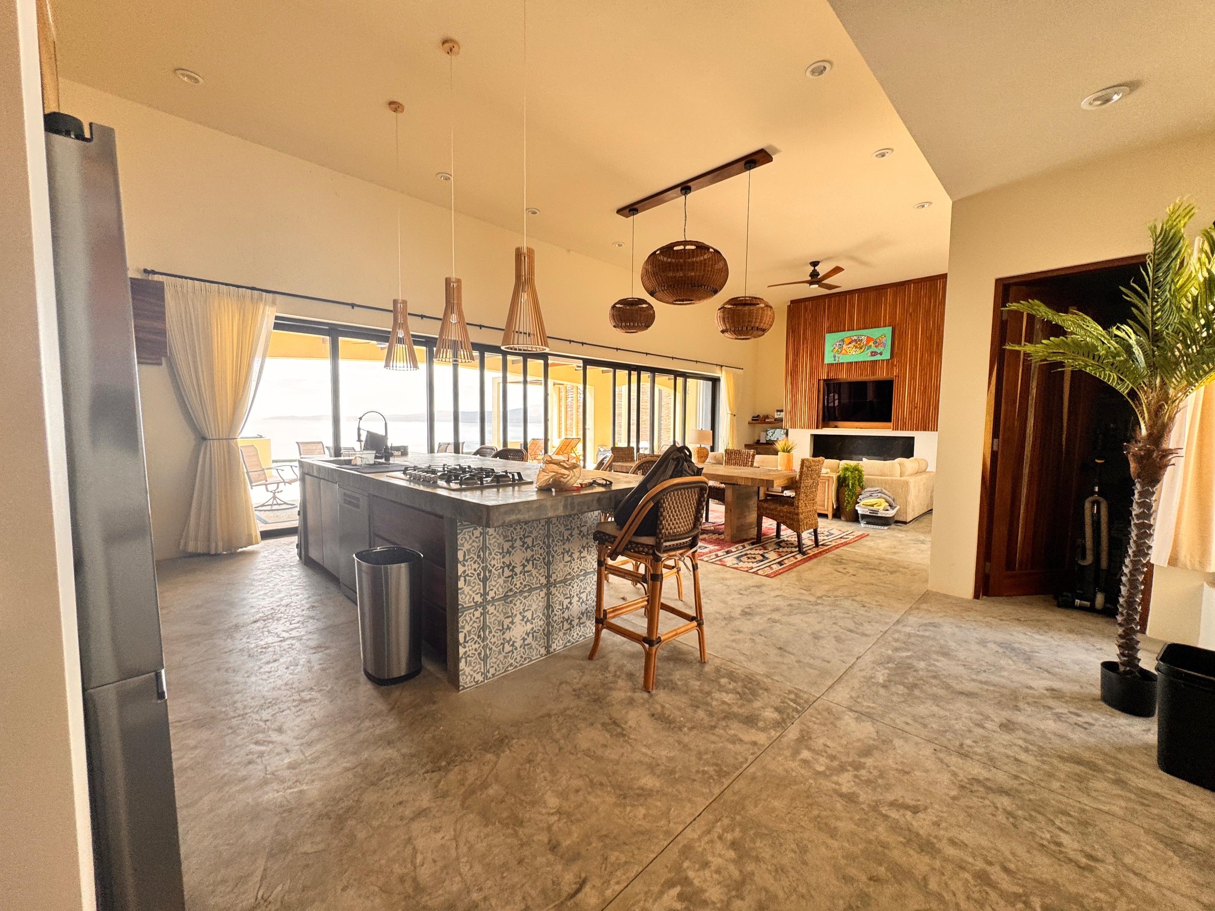 Open-concept living room and kitchen with large windows, beige sofas, a wooden entertainment center, and a kitchen island with hanging pendant lights.