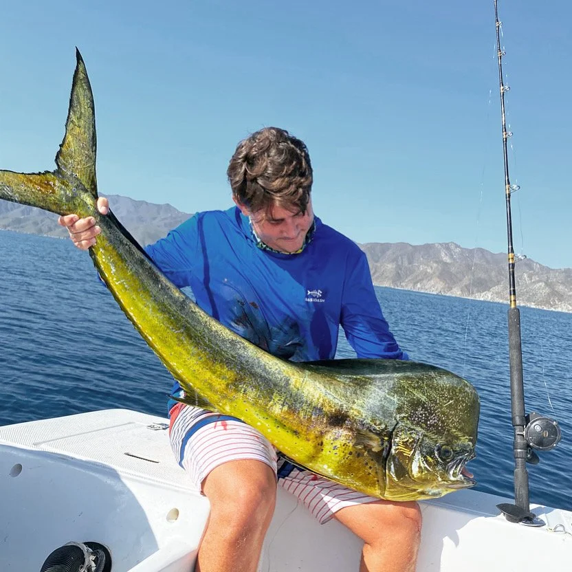 A young man in a blue long-sleeve shirt and striped shorts sitting on a boat holding a large fish, a mahi-mahi, with a fishing rod nearby, with water and mountains in the background.