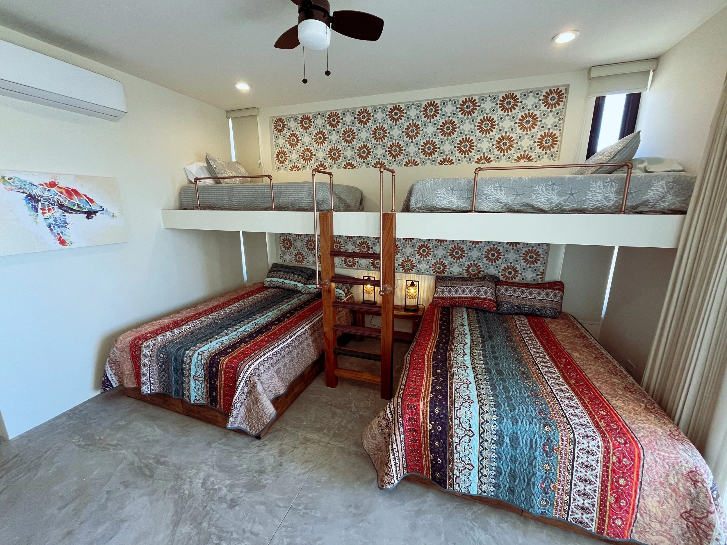 A bedroom with a bunk bed setup; two beds are on the floor and two beds are on an elevated loft, accessible by a ladder, with lamps and pillows. The room is decorated with colorful patterned bedding and wall art.