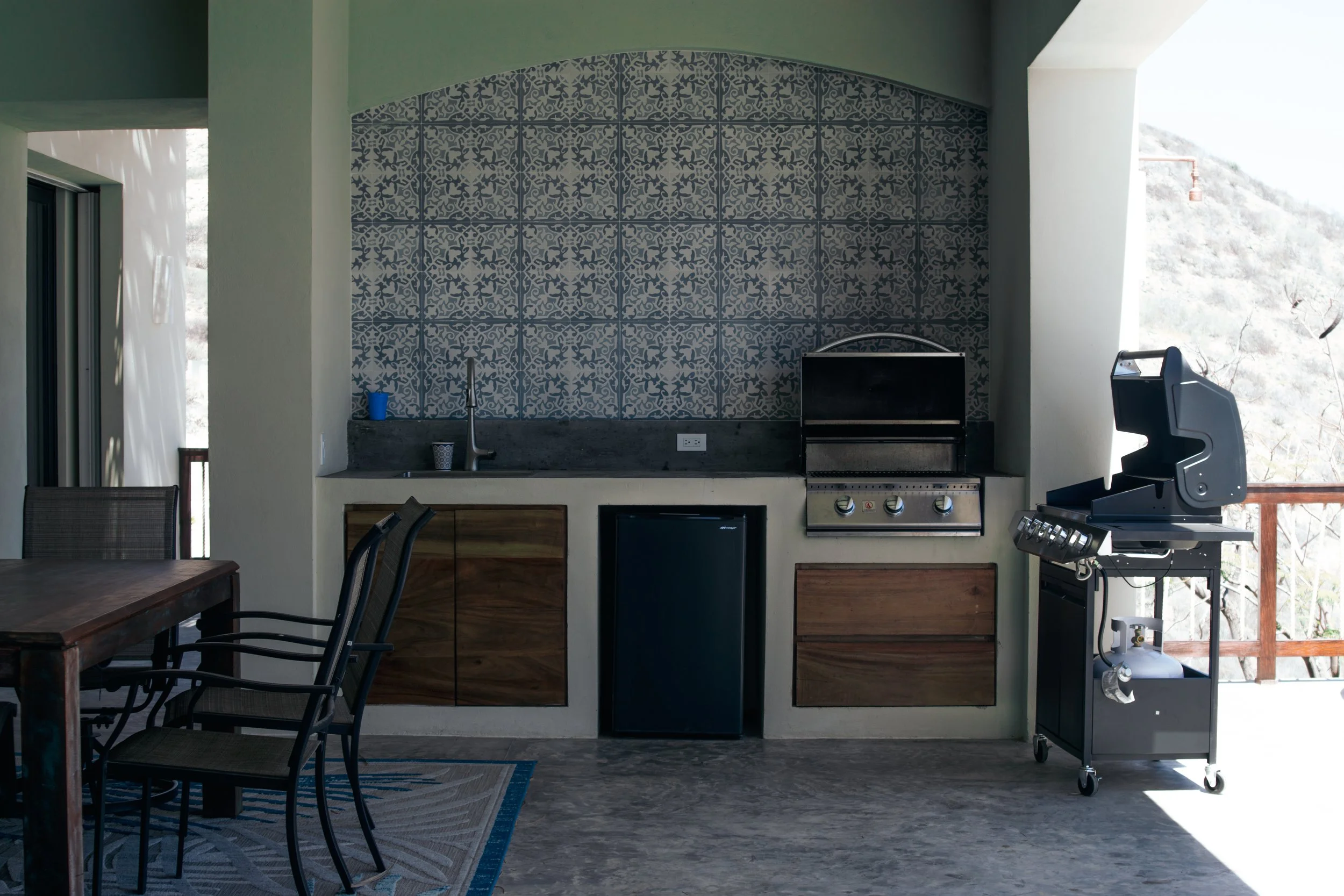 Outdoor kitchen area with a built-in grill, mini fridge, and a sink, adjacent to a balcony with a scenic mountain view.
