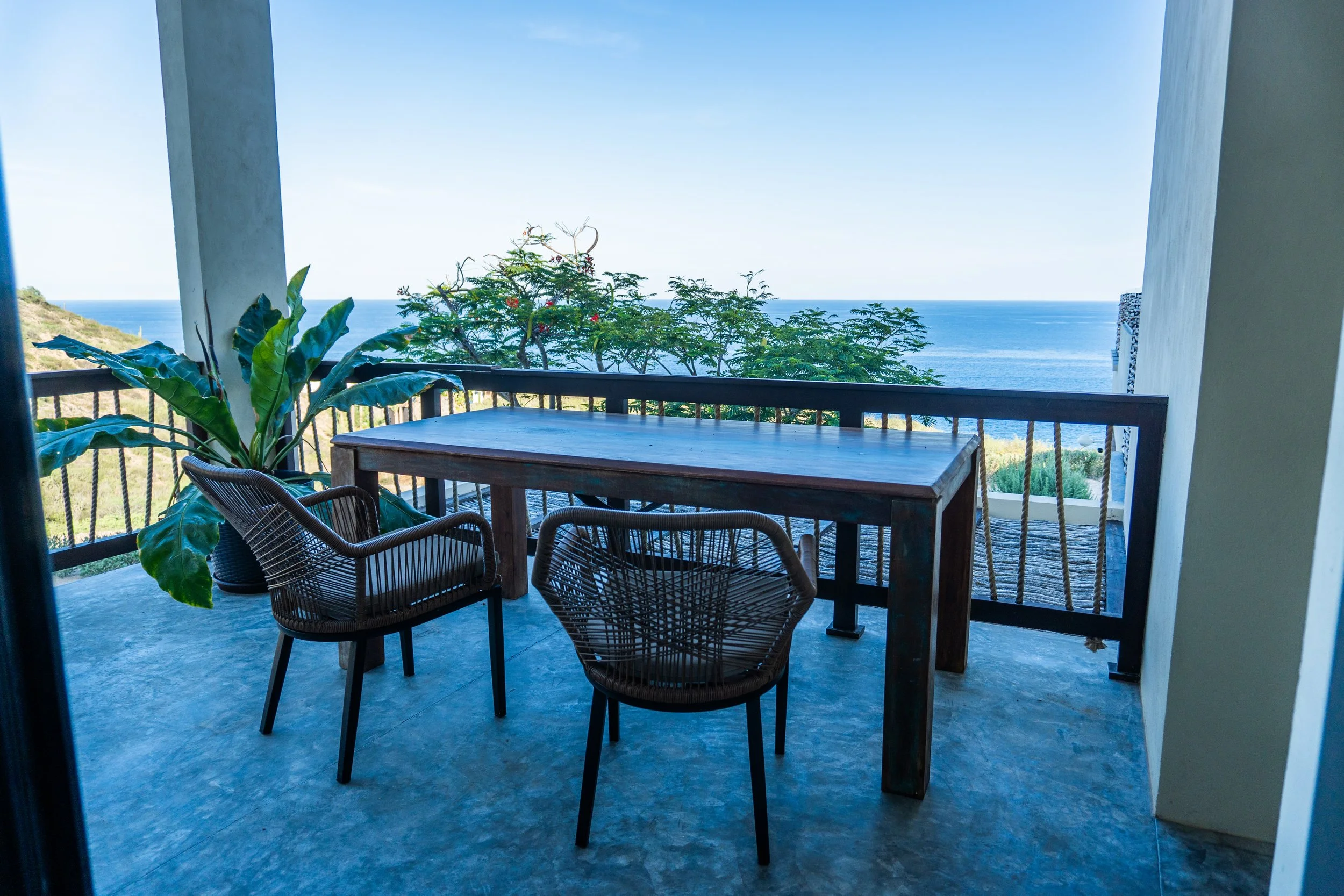 Balcony with wooden table and wicker chairs overlooking the ocean, with green foliage and a clear blue sky in the background.