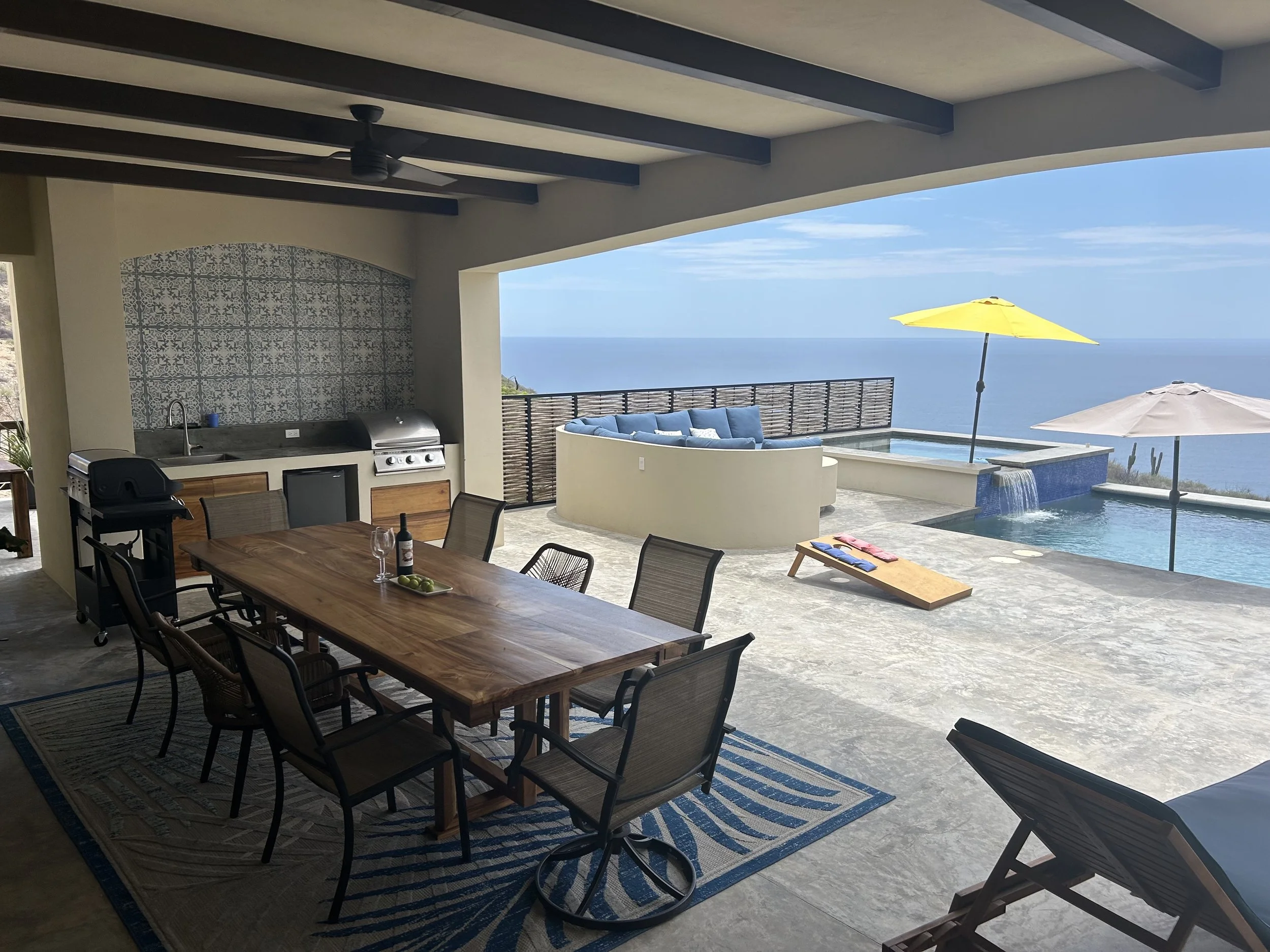 Outdoor patio with a dining table, chairs, a barbecue grill, a scenic view of the ocean, a pool with yellow and beige umbrellas, and lounge chairs.