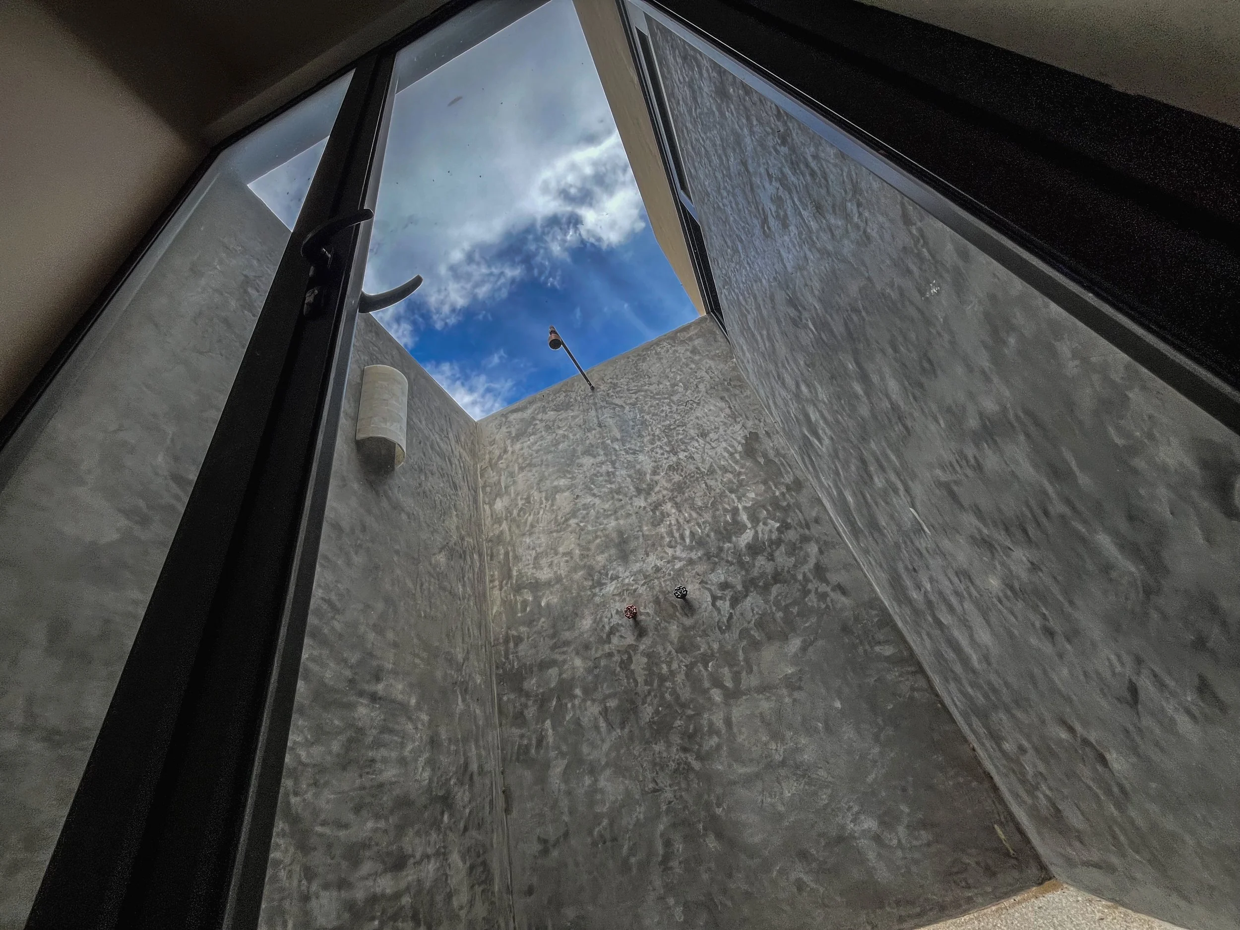View from inside a building looking up through an open rectangular skylight to a partly cloudy sky. The interior walls are textured and gray.