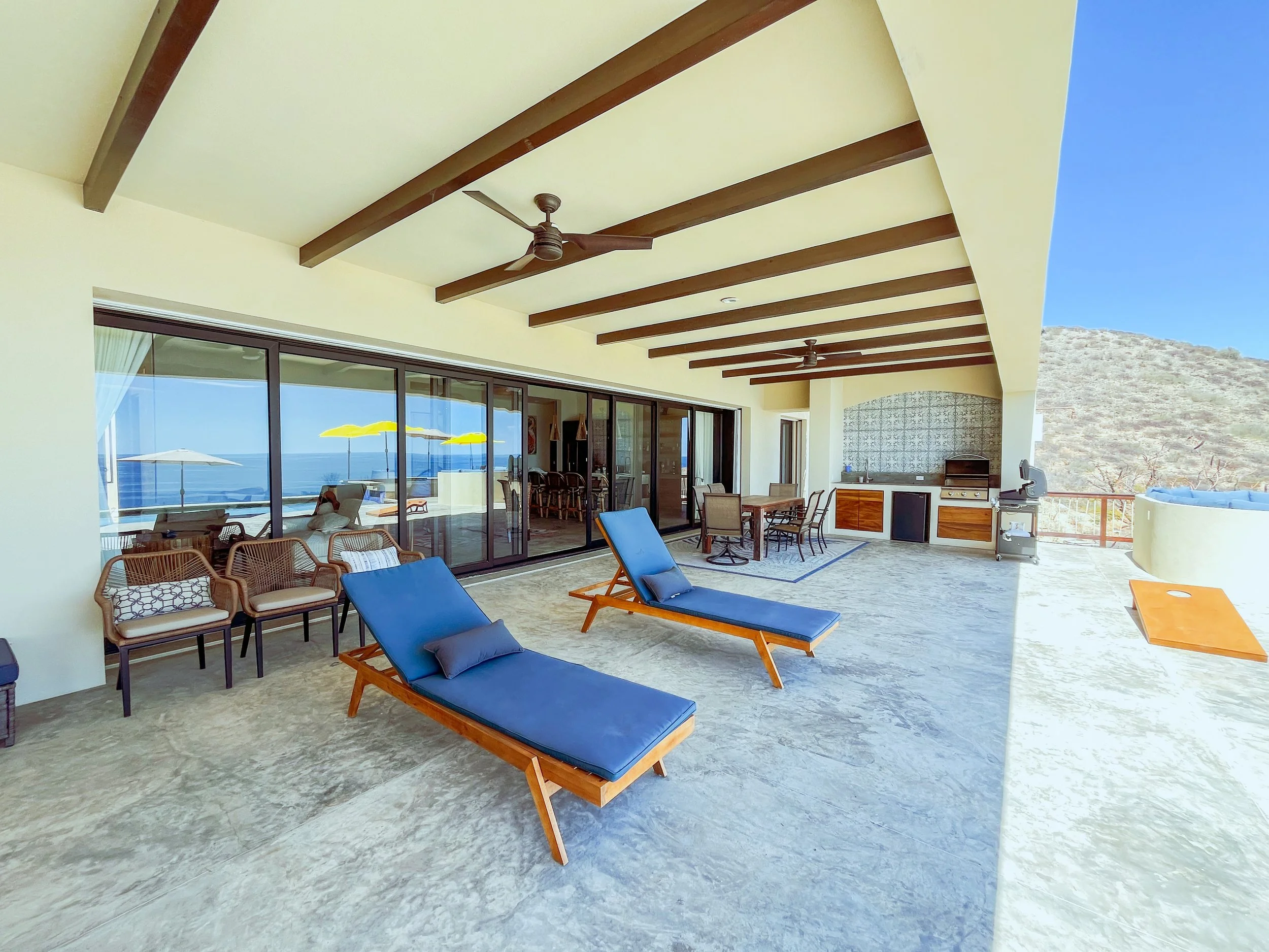 Spacious outdoor balcony with lounge chairs, dining area, and outdoor kitchen, overlooking a desert landscape with a mountain in the background.