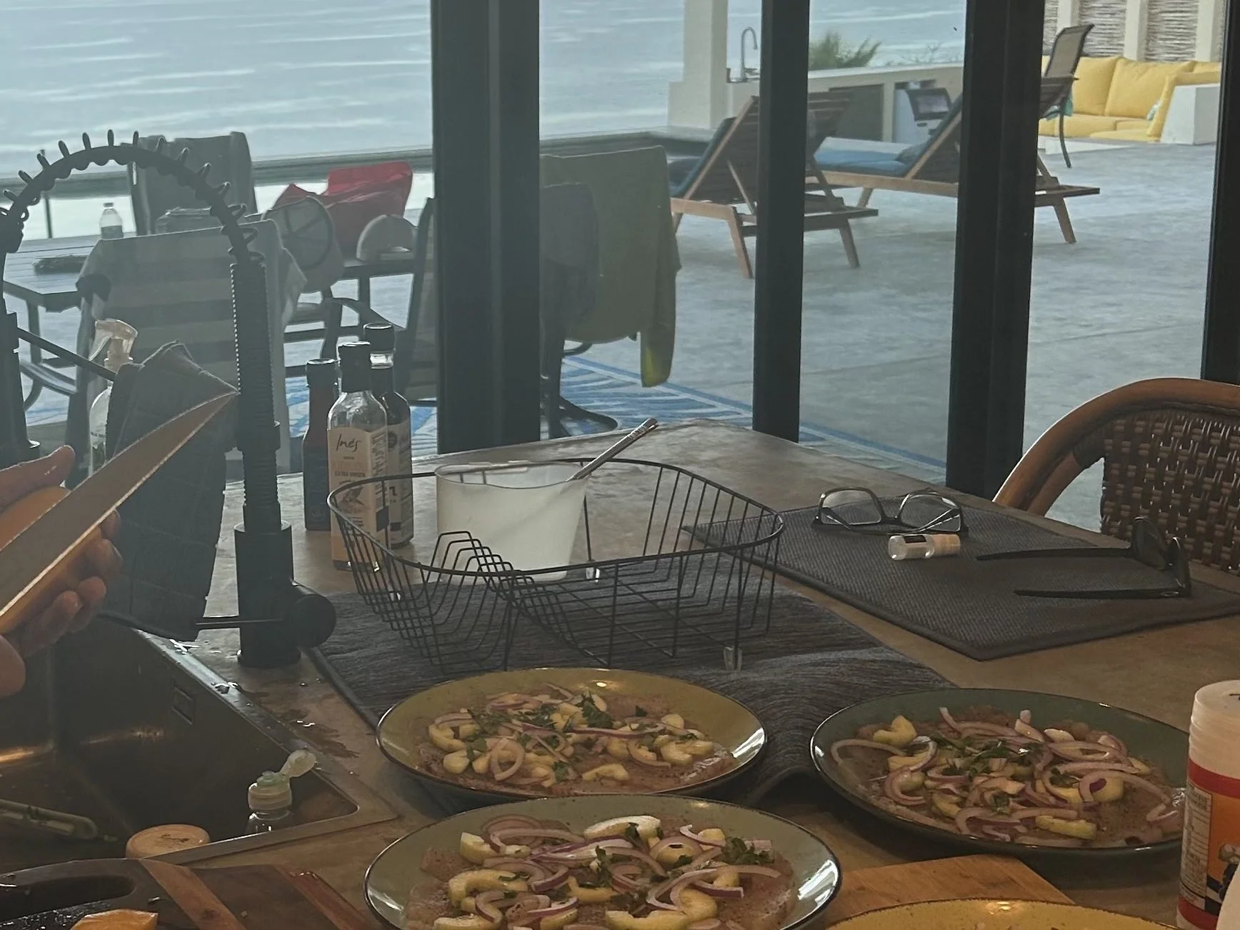 Two plates of pizza topped with onions and herbs on a kitchen counter, with a sink, dish soap, and a person holding a phone in the background, and a glass of ice or a drink in a bowl.